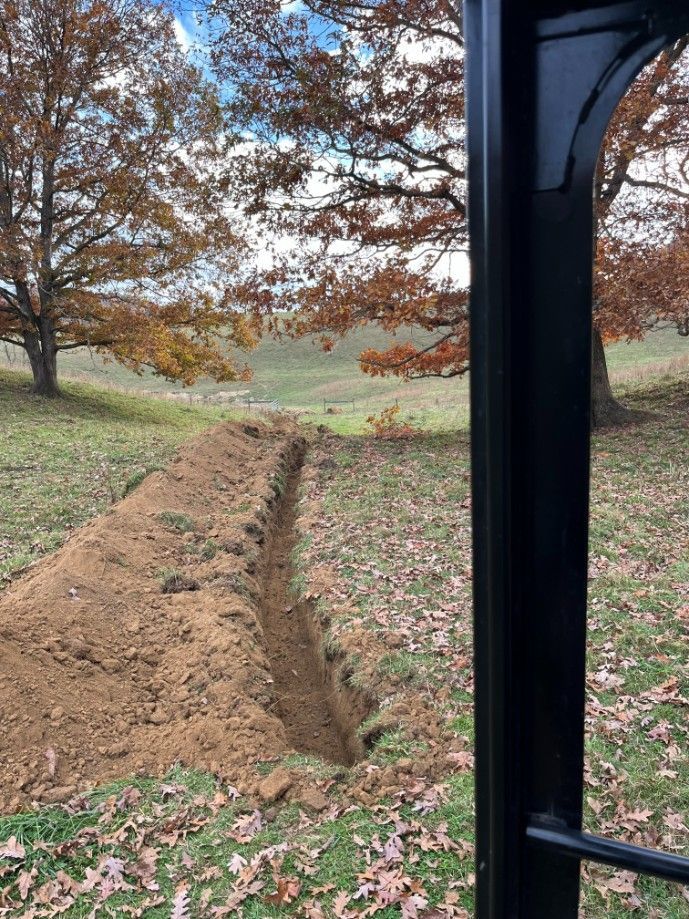 A long trench dug in a grassy field, viewed from behind a black fence post; trees with fall foliage in the background.