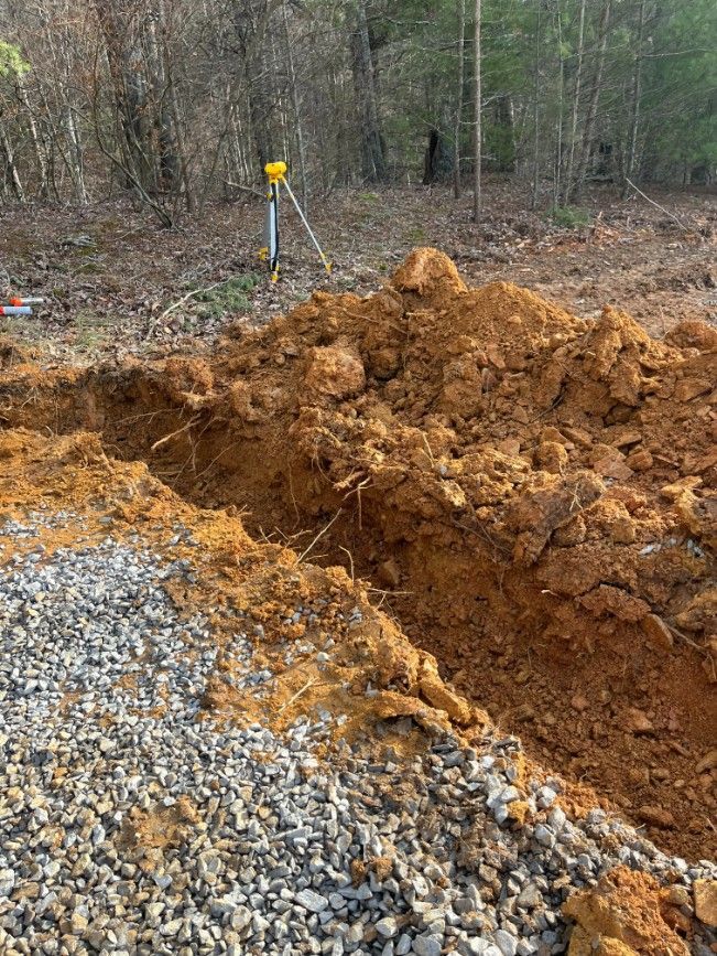 Dug trench with dirt pile and surveying equipment on a gravel path near trees.
