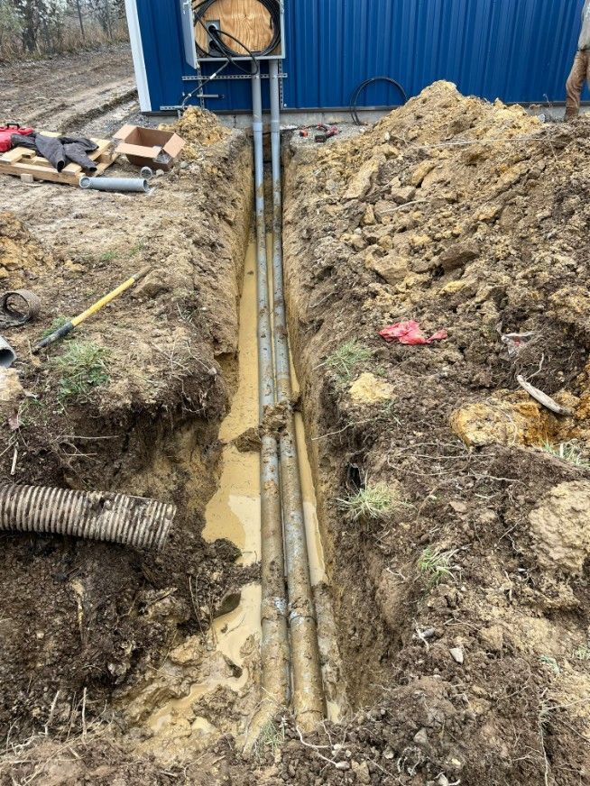 Trench with buried conduit near a blue building. Earth-toned mud and utility lines are visible.