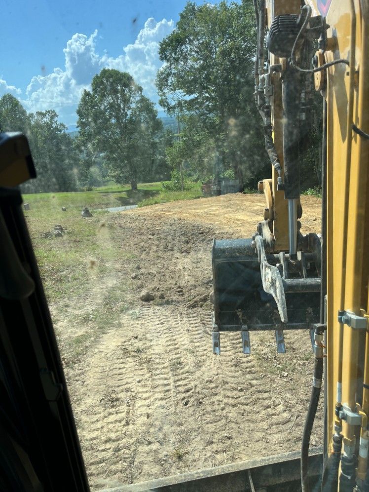 View from excavator cab; bucket and work area outdoors, with trees and cloudy sky in background.