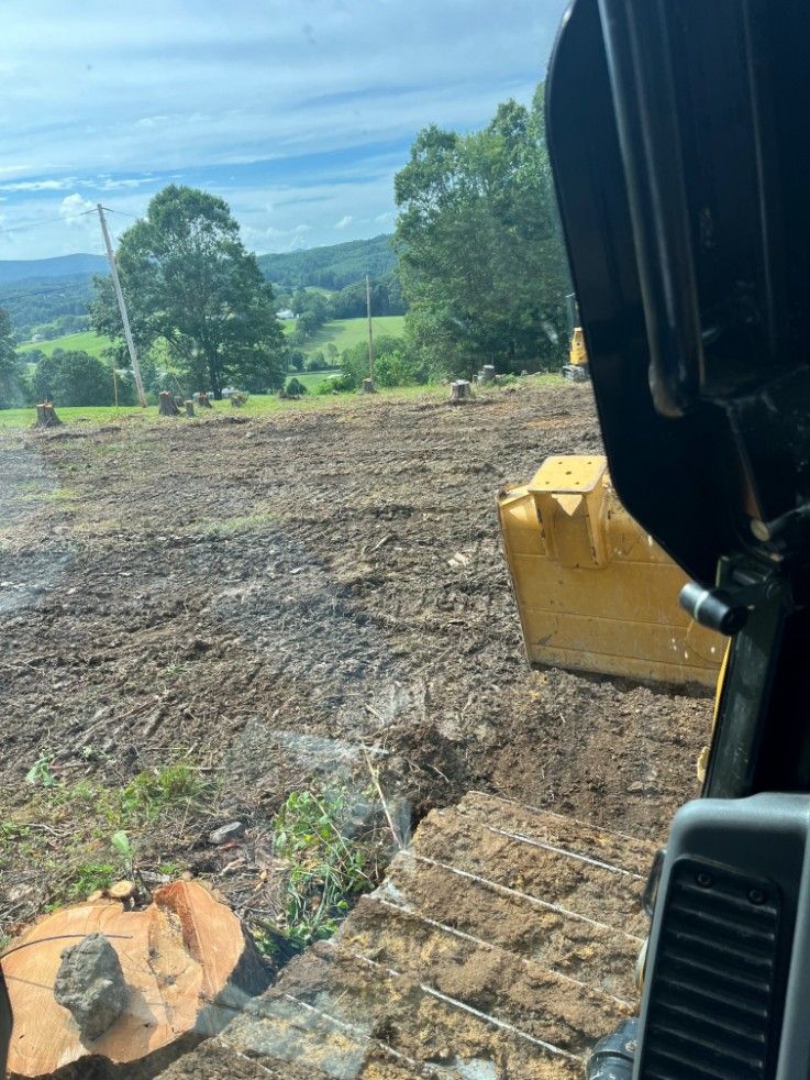 View from inside a bulldozer. Clearing land with dirt, trees, and a distant green landscape.