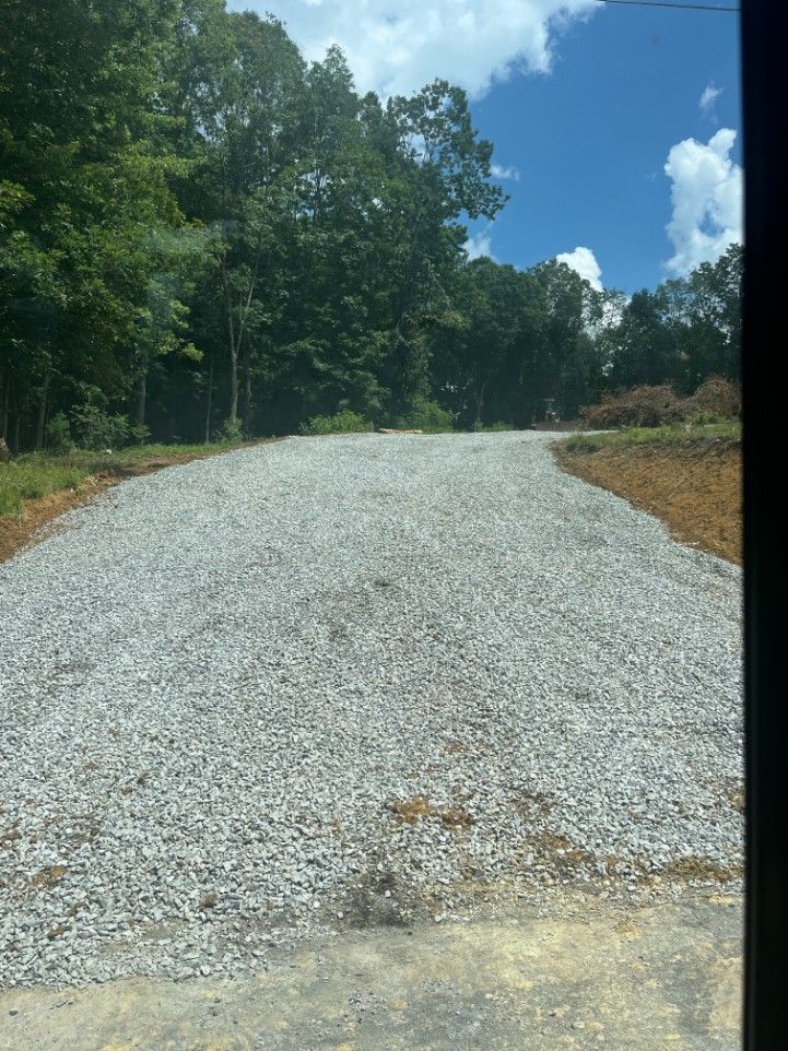 Gravel driveway leading uphill towards trees under a partly cloudy blue sky.