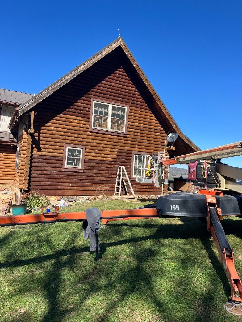 Brown log cabin with two white windows, ladder, and a boom lift on a green lawn against a clear blue sky.