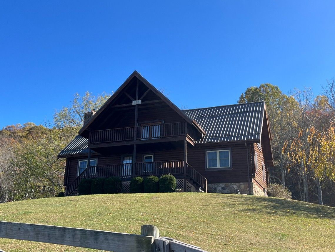Brown cabin with a black roof on a grassy hill, surrounded by trees under a clear blue sky.