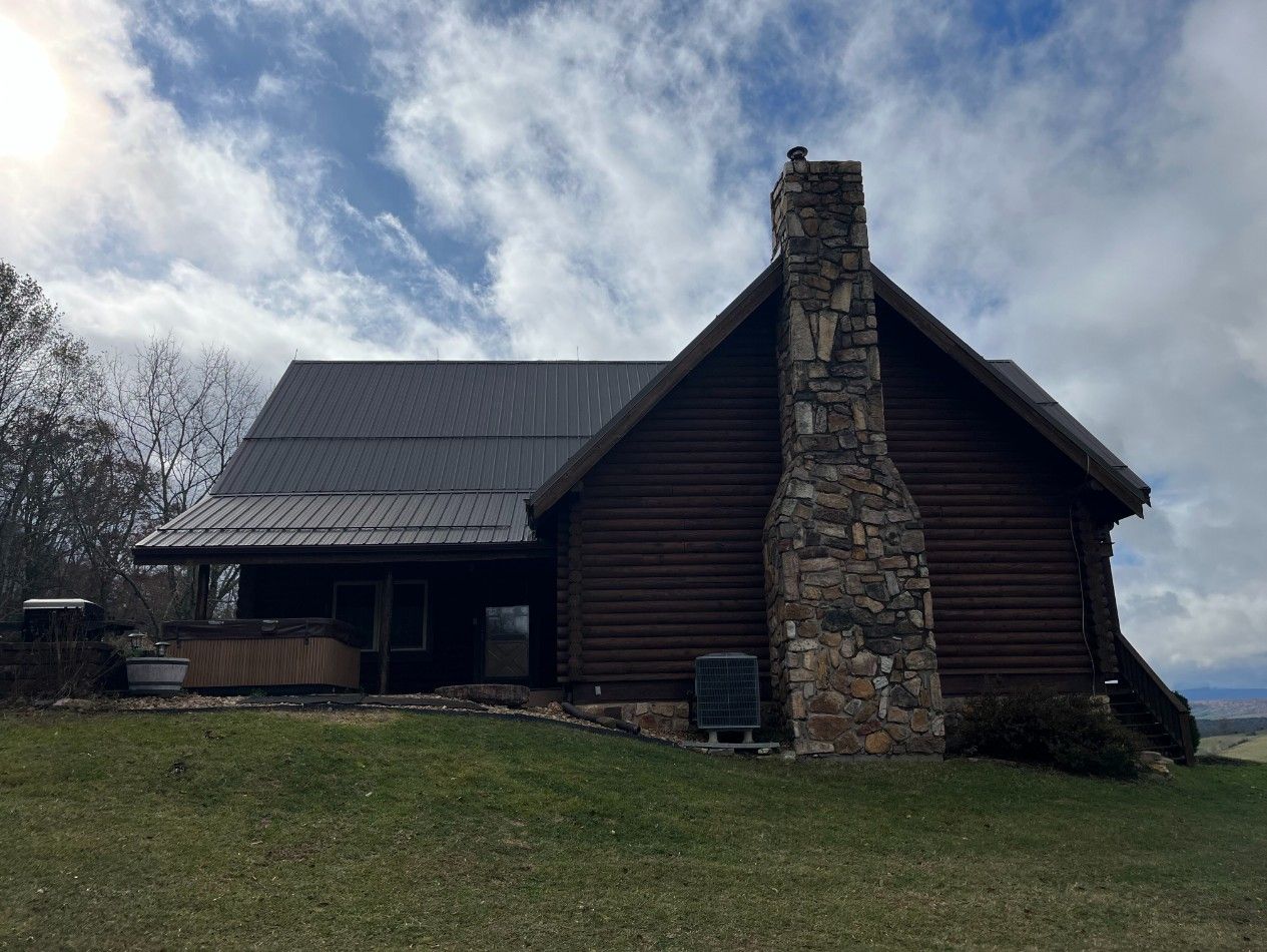 Log cabin with stone chimney, dark roof, and covered porch on a grassy hill under a cloudy sky.