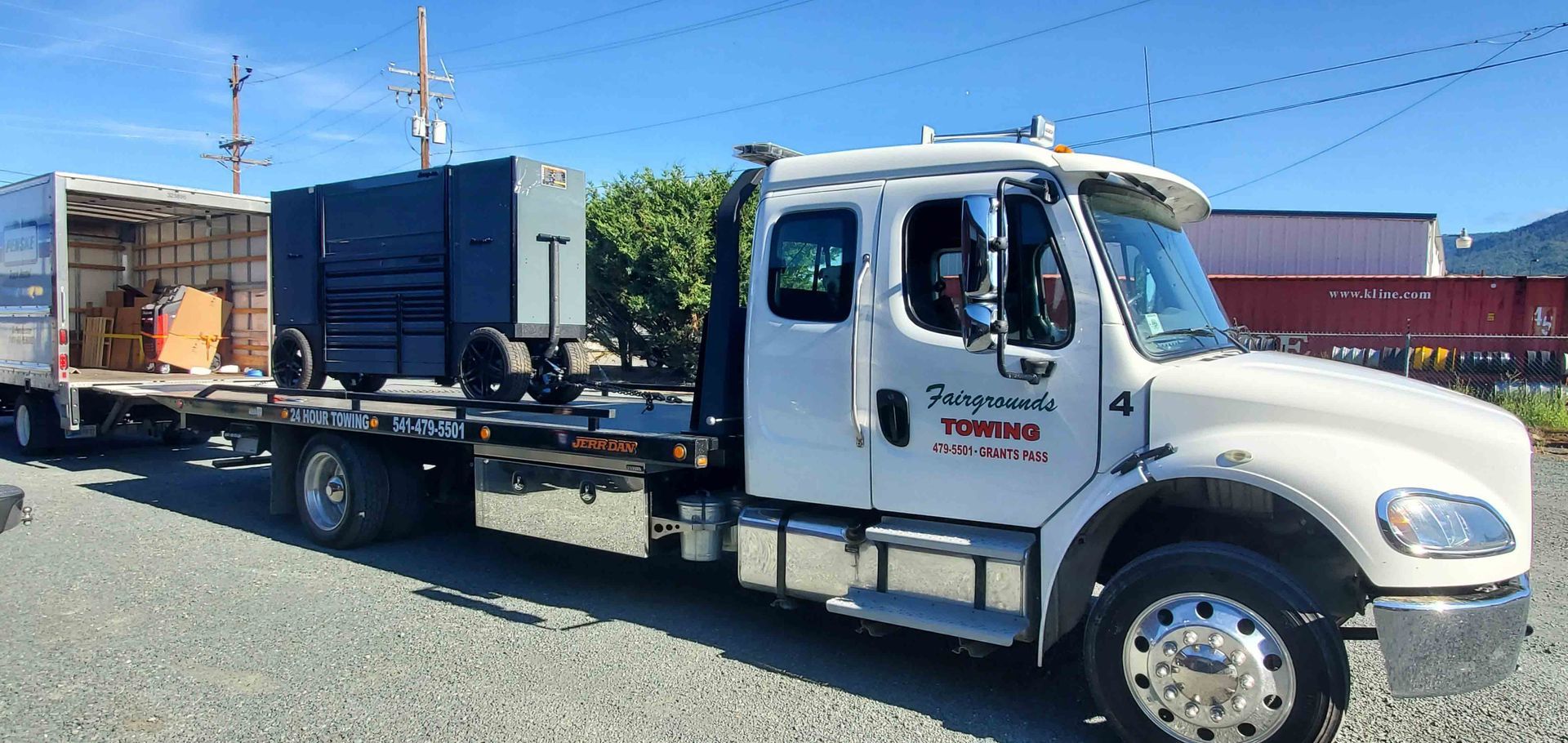 White flatbed tow truck carrying a blue dumpster container in a gravel lot under a clear blue sky