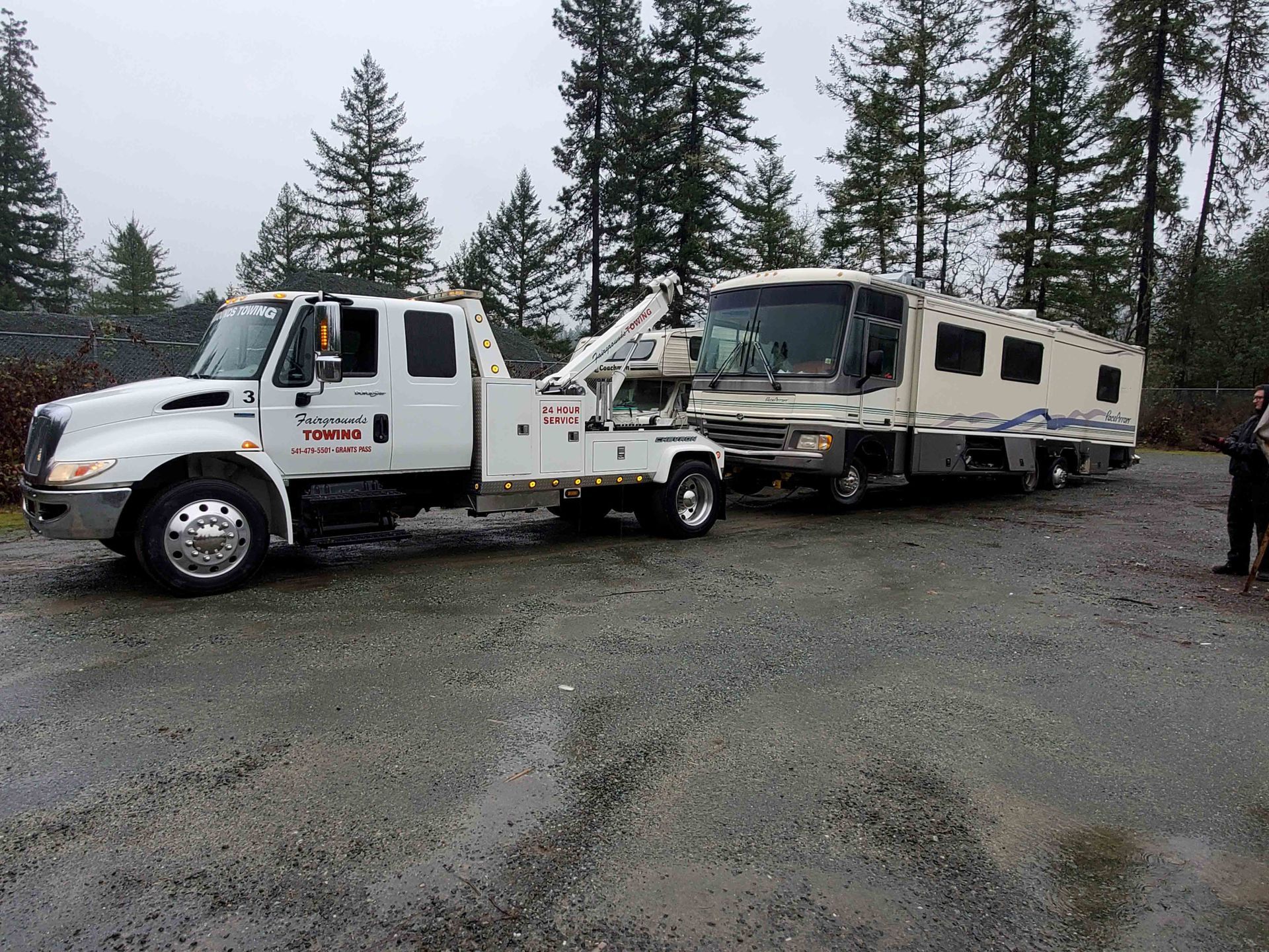 Tow truck hauling a large RV on a gravel lot with pine trees in the background