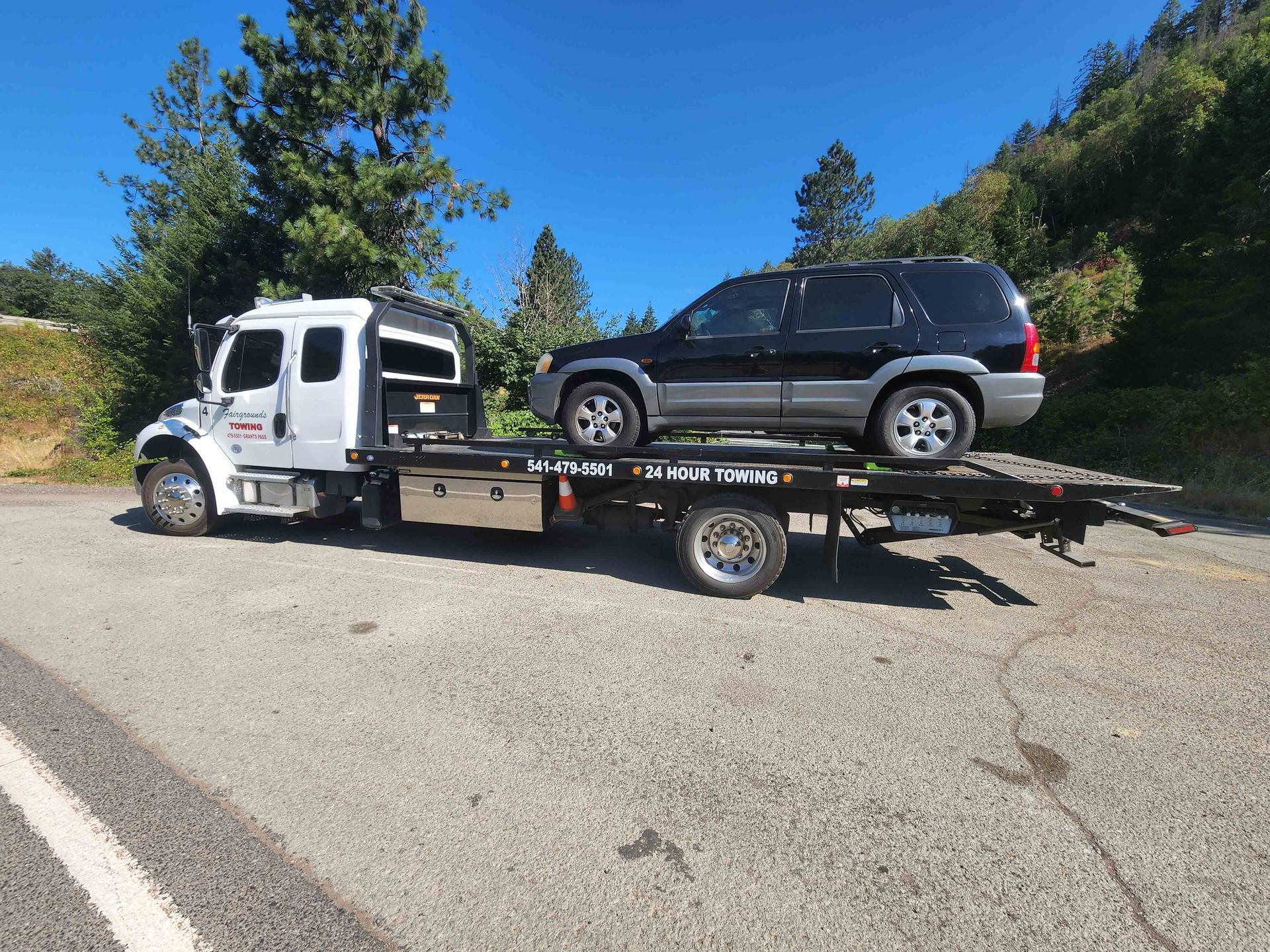 White flatbed tow truck carrying a black SUV on a sunny roadside with trees and hills.