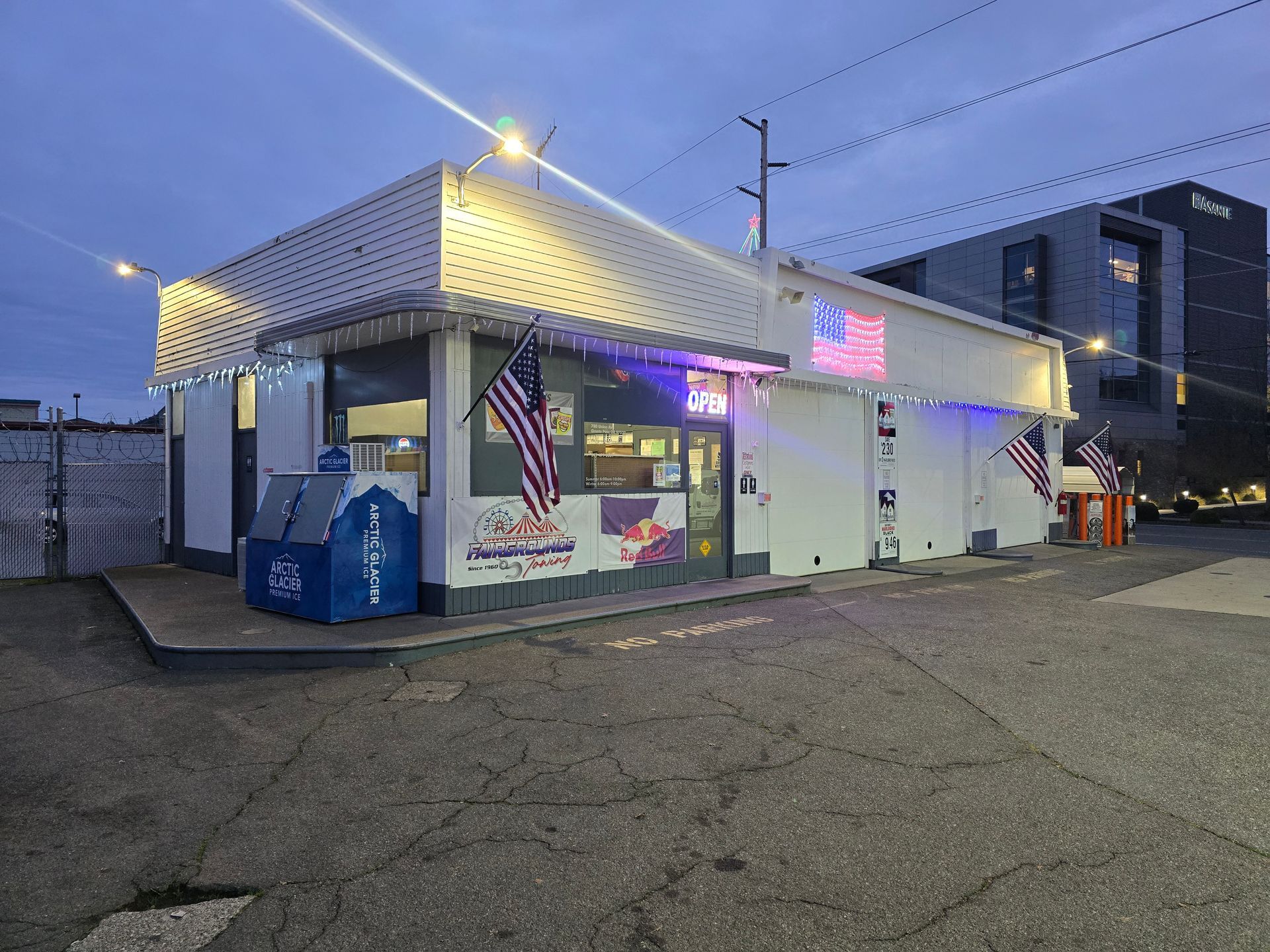Modern roadside gas station services with red canopy and fuel pumps under blue sky. Small white storefront at dusk with lit awning, neon sign, and parked cars in a lot