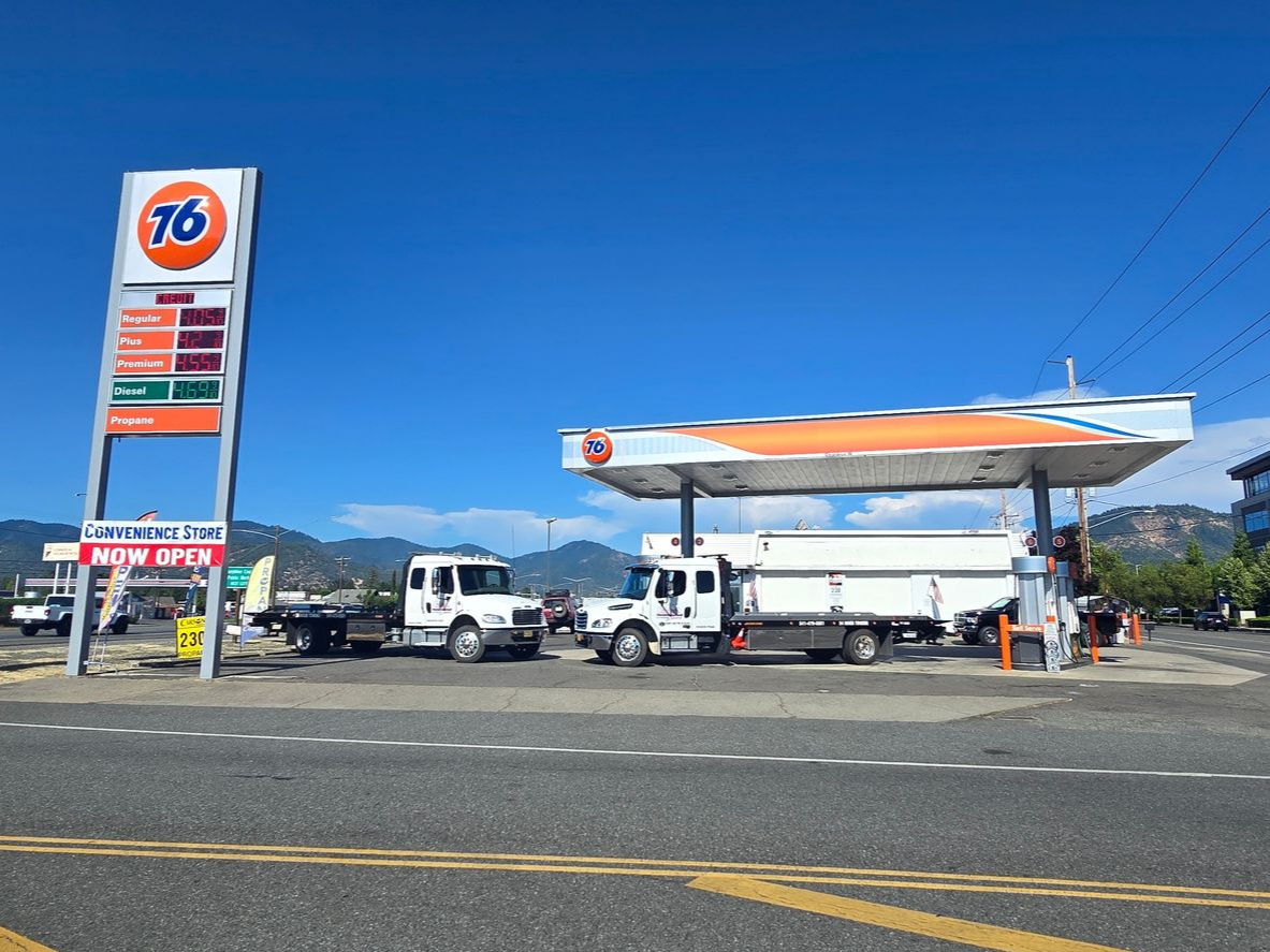 A Man Kneeling Next To Two Dogs In Front Of A Tow Truck — Grants Pass, OR — Fairgrounds Towing & Fuel LLC 76 gas station with white trucks under an orange canopy on a sunny roadside