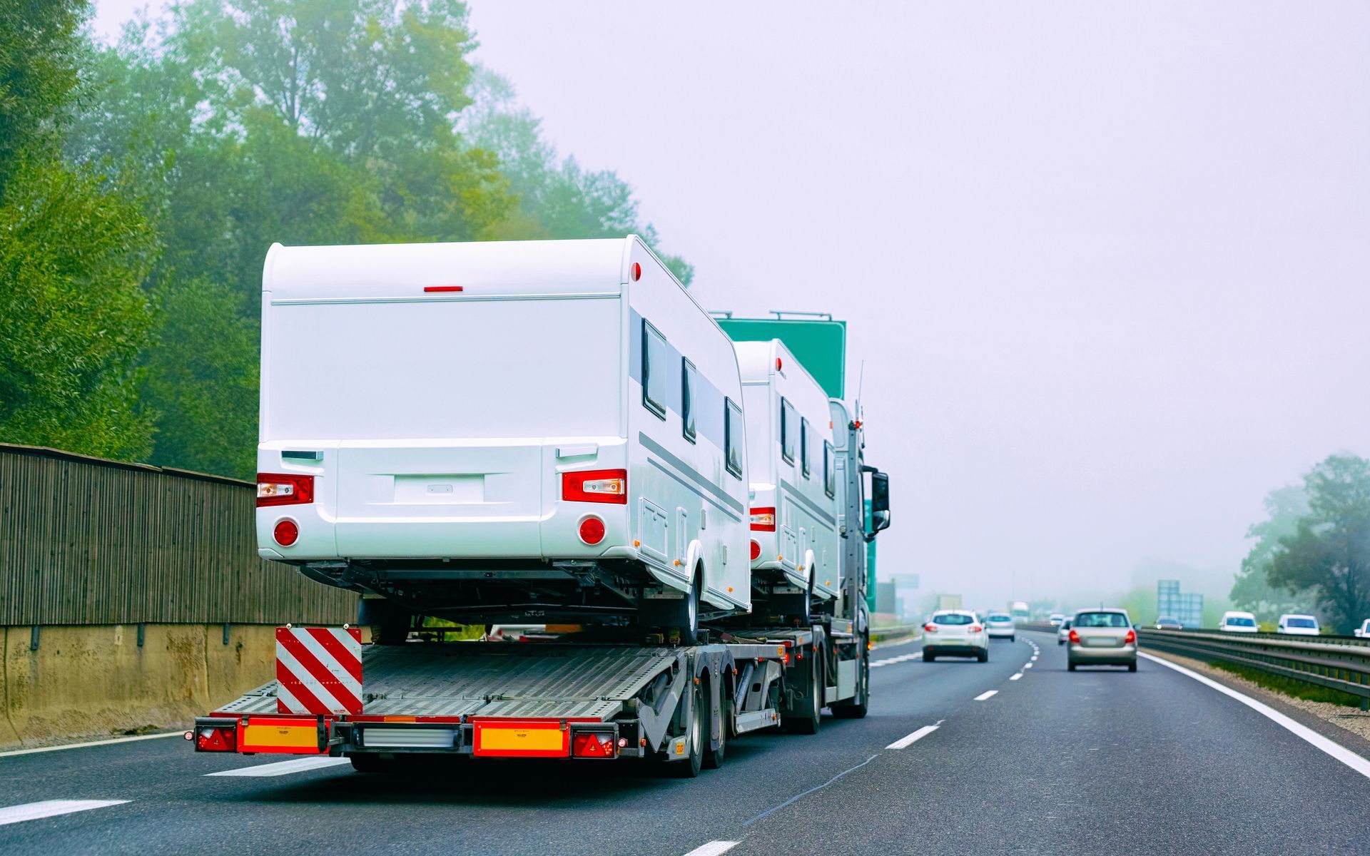 Semi-truck representing safe recreational vehicle towing hauling travel trailers on busy highway. Flatbed truck carrying white caravans on a wet highway, with cars ahead and trees along the roadside.