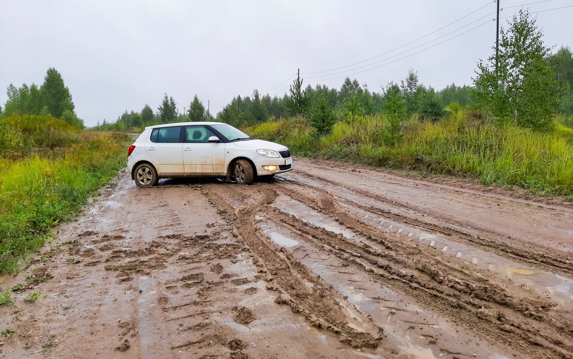 A white passenger hatchback stuck on a muddy road in rainy weather. White car stuck in muddy ruts on a dirt road, surrounded by green shrubs and trees