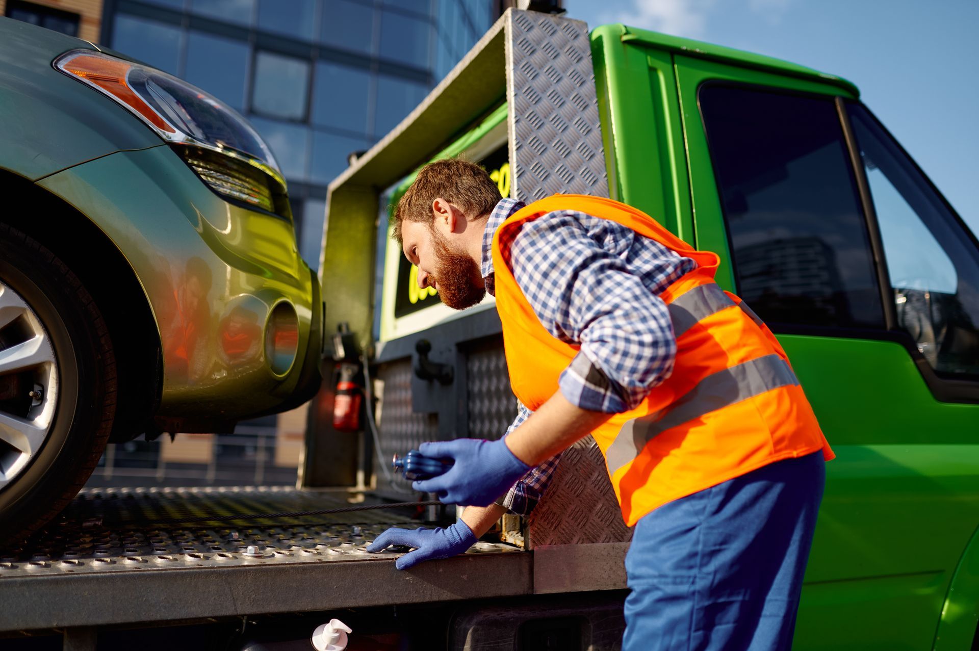 A man working in a towing service on a road. Mechanic in orange safety vest working on car being loaded onto a green tow truck