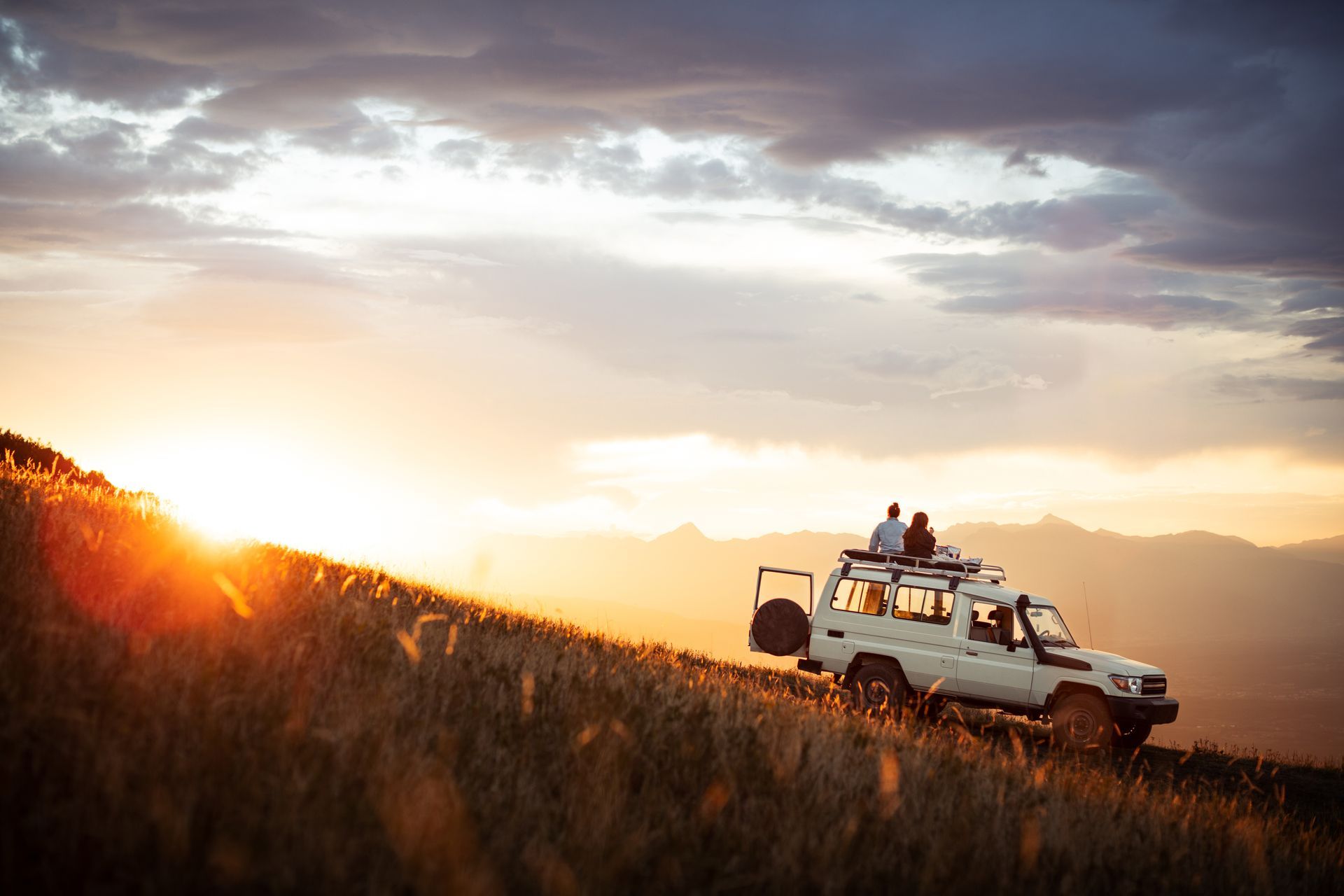 Two people watching a sunset on top of a 4x4 vehicle overlooking mountains.