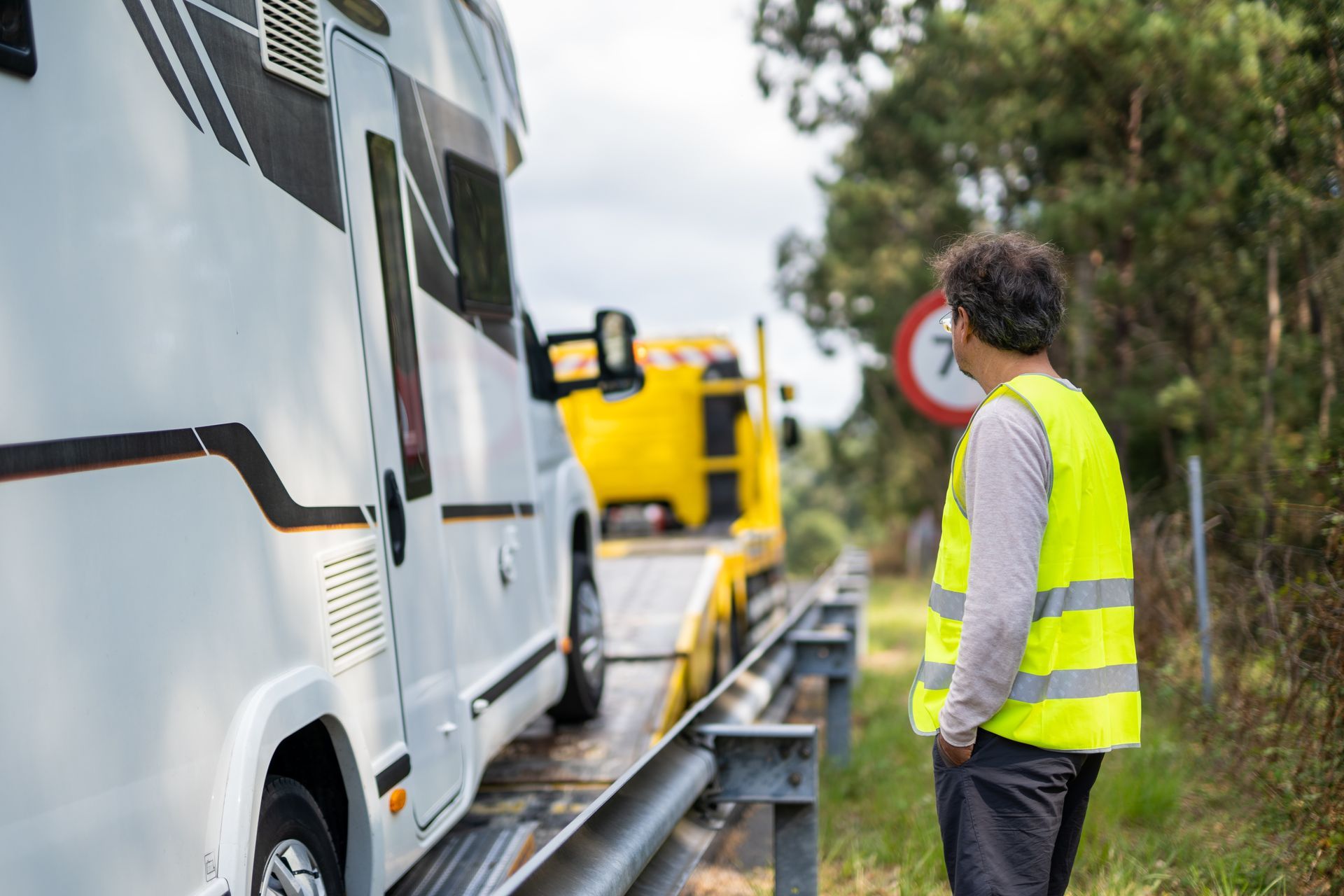 A man standing next to a motor home being towed Tow truck loading a white van roadside, with a worker in a neon yellow safety vest nearby