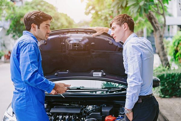 Two men talk beside an open car hood in a driveway, examining the engine.