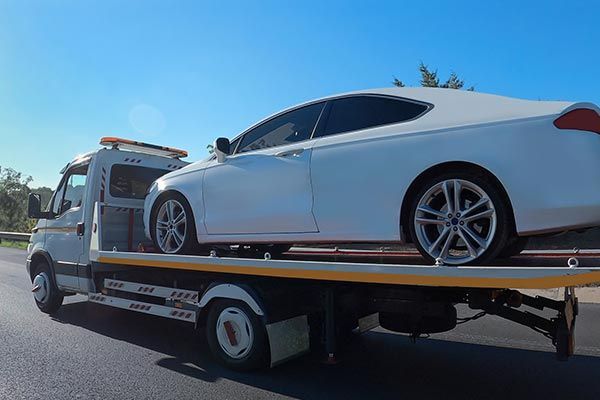 White sedan on a tow truck, driving on a sunny road.
