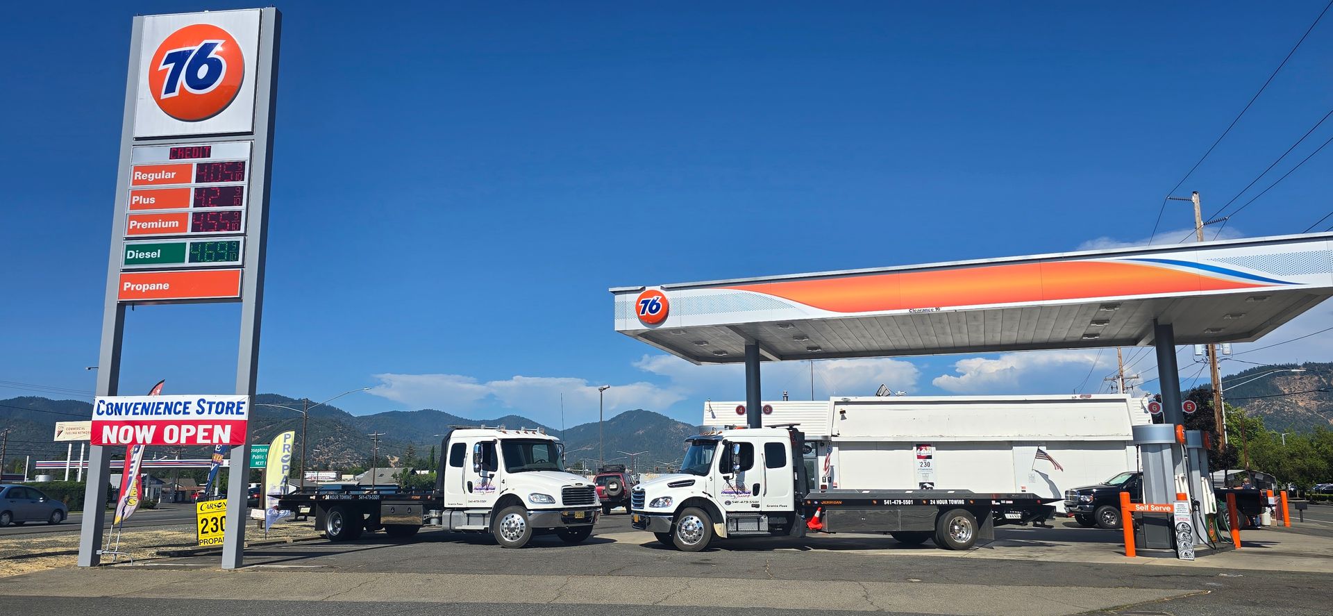 A man working on a towing service on a road. 76 gas station with fuel trucks parked under an orange-and-white canopy on a sunny day