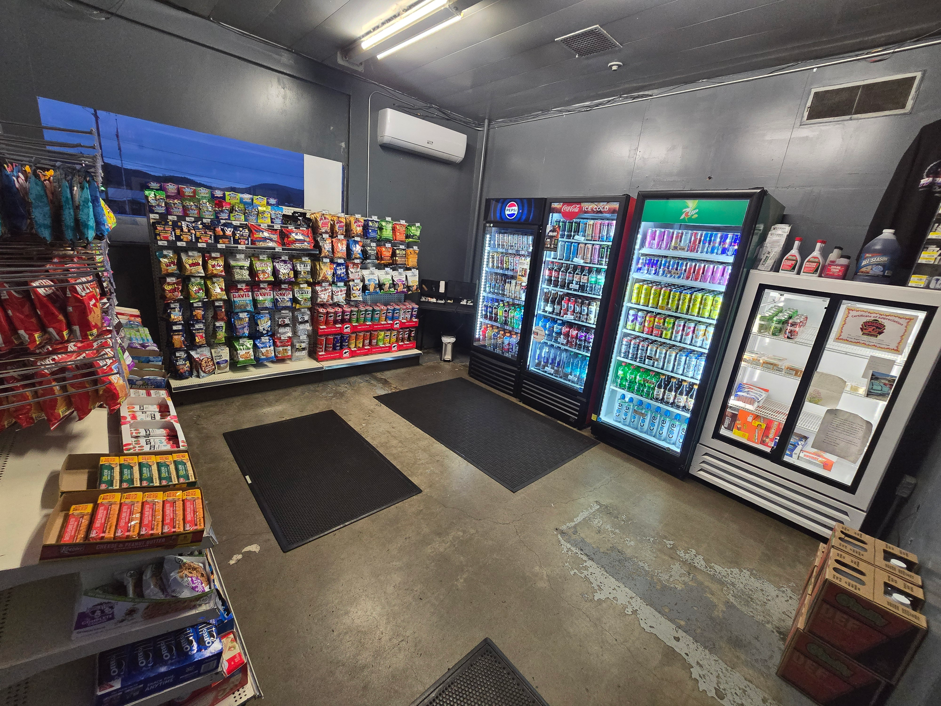 Small convenience store interior with snack shelves, coolers, and a few floor mats on a gray floor.