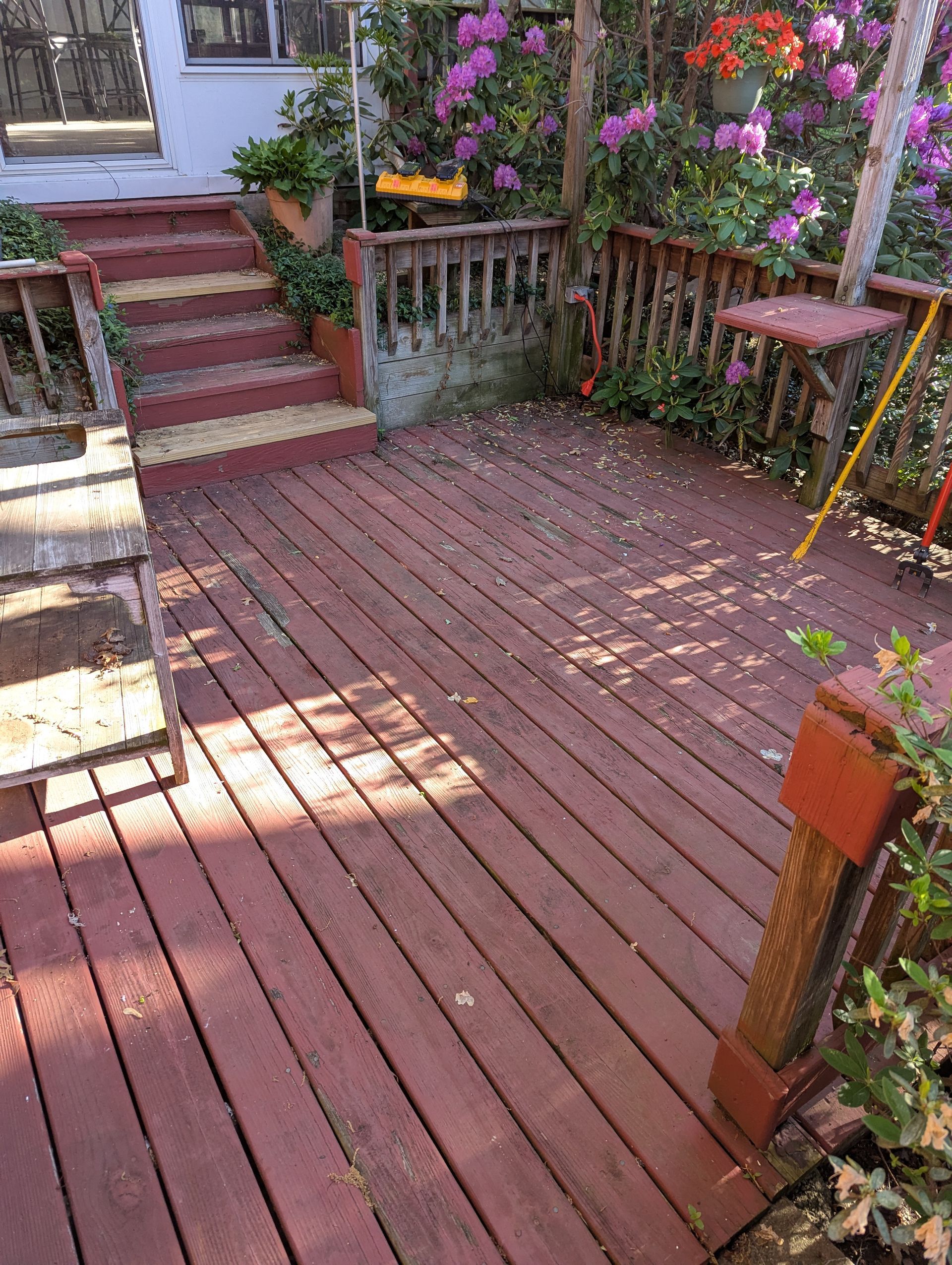 Red wooden deck with steps, railing, and potted flowers in a sunny backyard garden