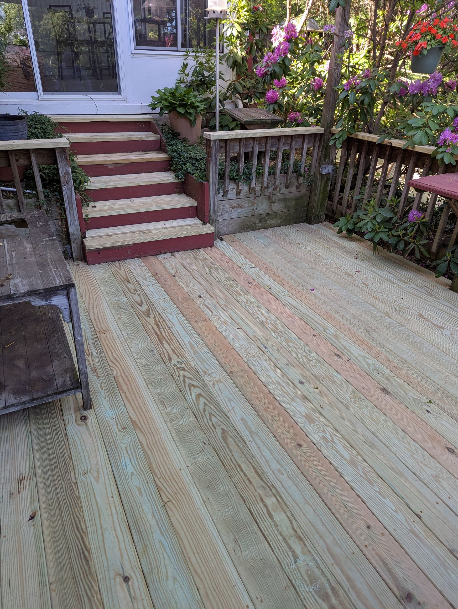 Backyard deck that has been sanded and repaired with red steps, wooden railing, beside flowering plants.
