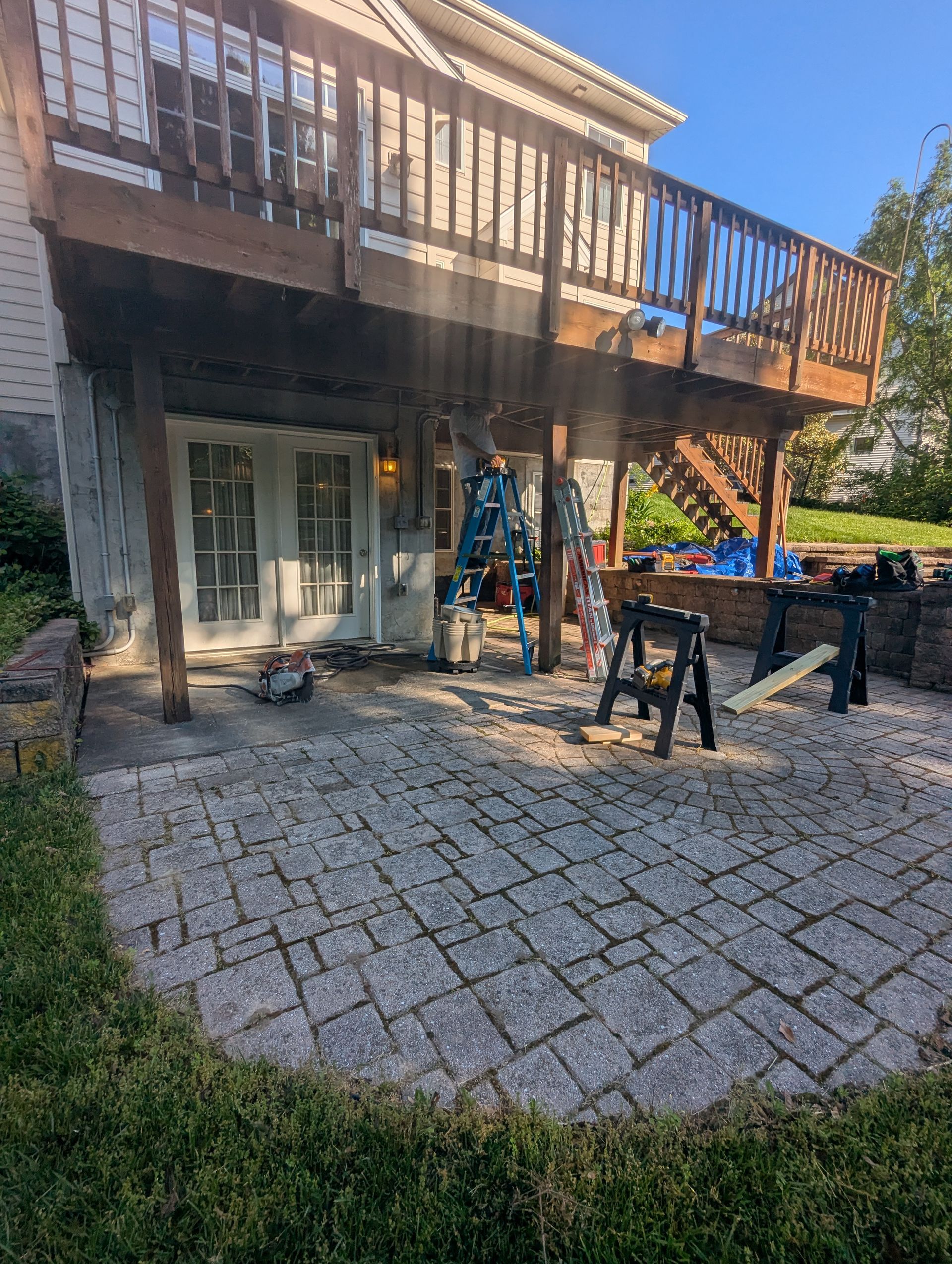 Backyard patio with a raised wooden deck, stone pavers, and construction tools under the deck