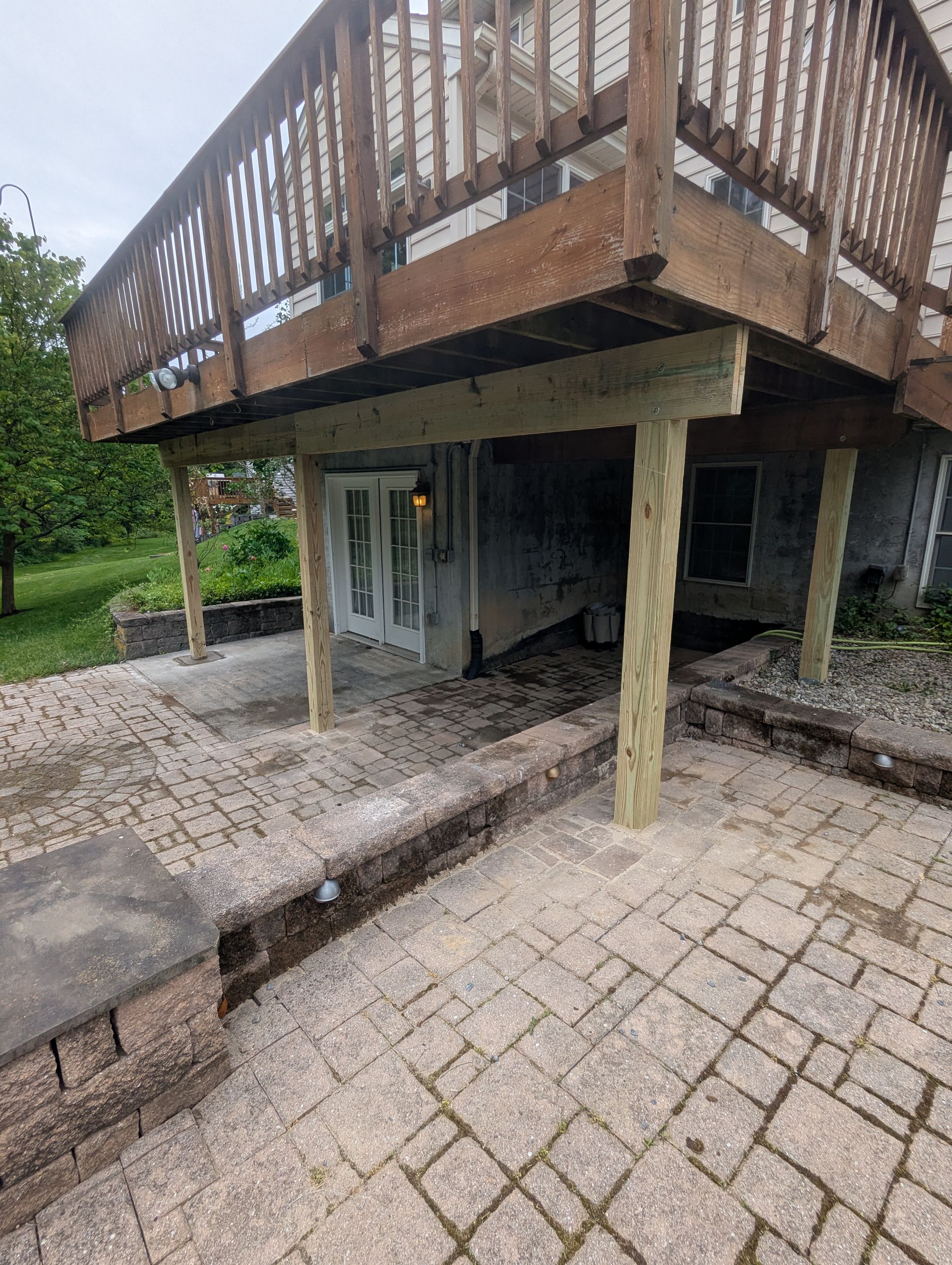 Covered wooden deck over a small patio and stair area beside a paved walkway in a yard.