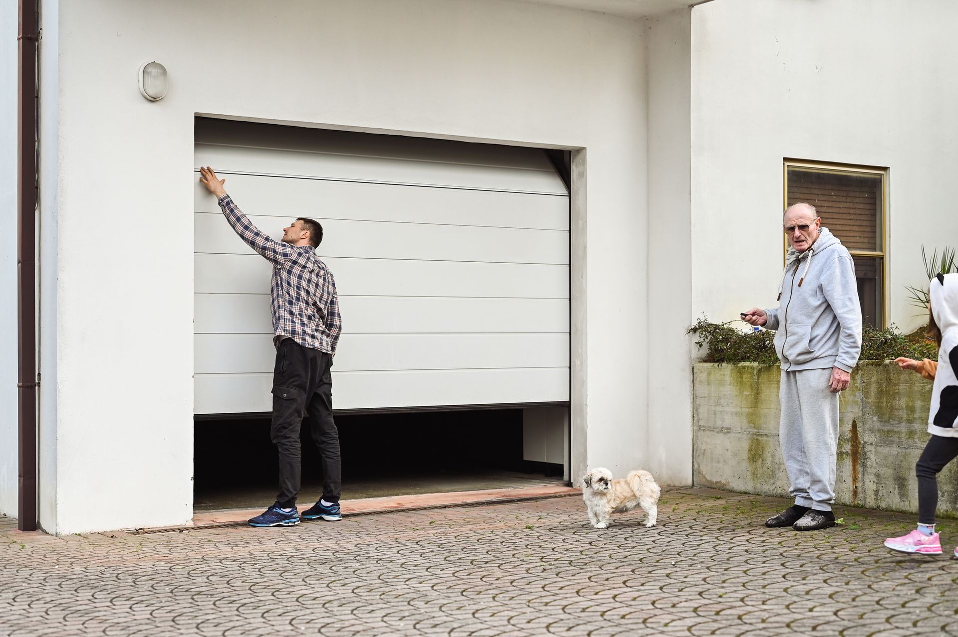 Man opening a white garage door, with a dog, older man, and child nearby on a paved driveway.