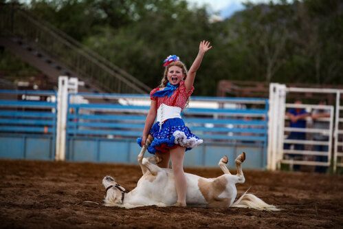 Cowgirl performer in blue dress posing on a dirt arena with two goats lying at her feet