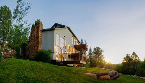 Modern white house with lit deck at sunset on a green hillside, trees and rocks in the foreground