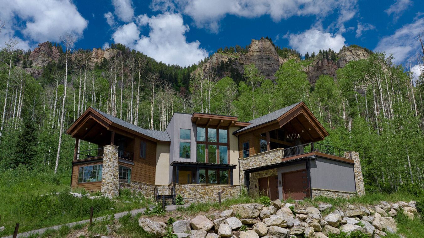 Modern mountain house with wooden roofs, stone walls, and large windows beneath rocky, forested cliffs.
