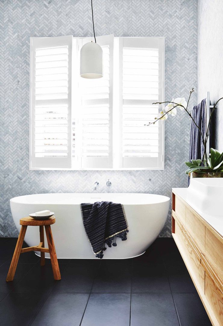 Bathroom with a white tub, herringbone tile wall, window with shutters, wooden stool, and black floor.