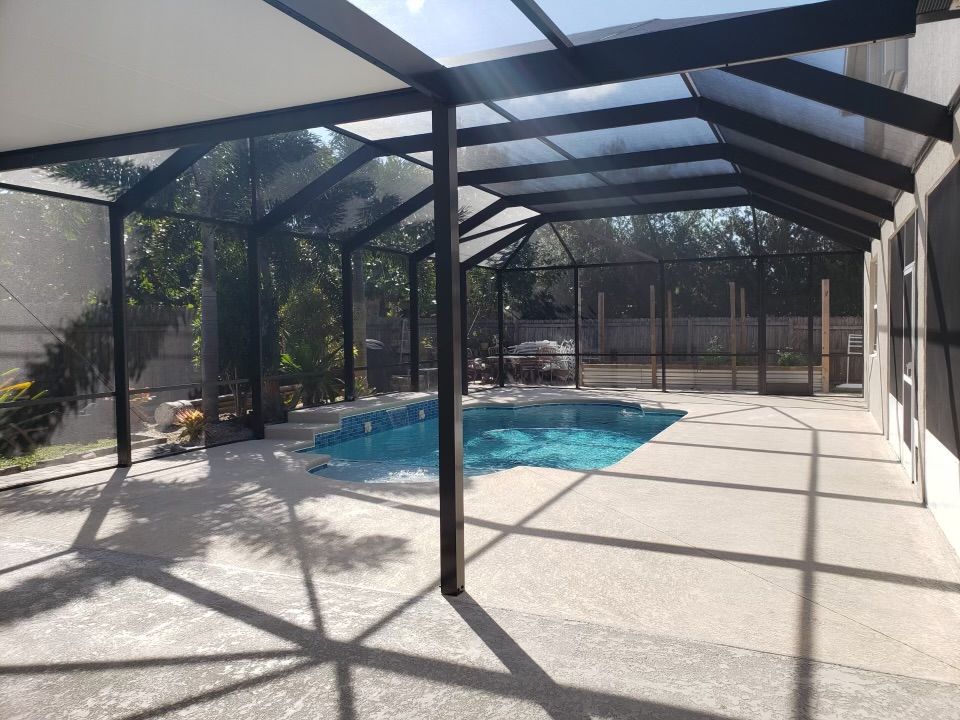 Pool enclosure with a swimming pool. Sunlight filters through the roof creating shadows on the concrete deck.