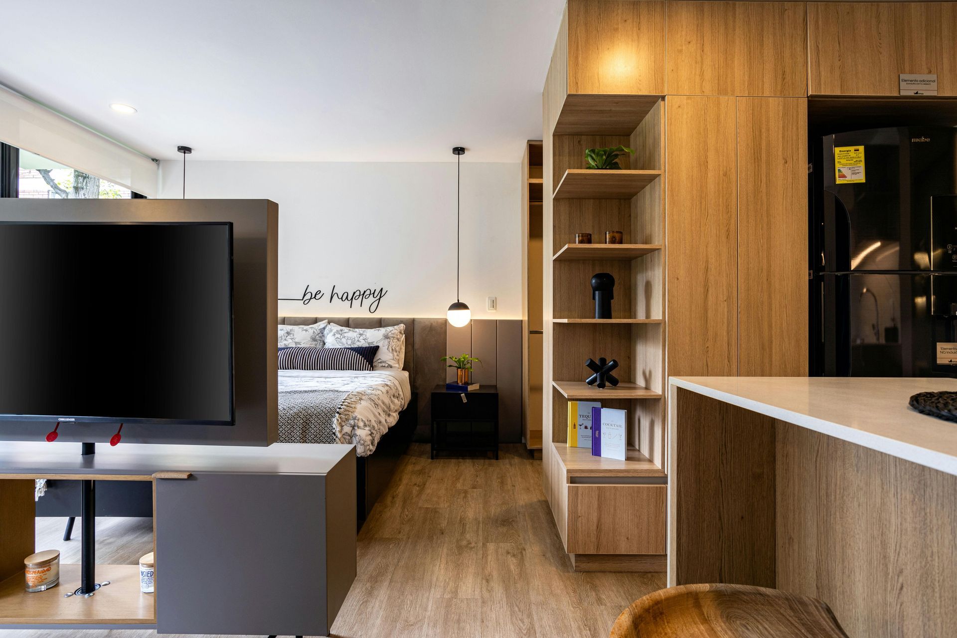 Modern studio bedroom with wood cabinetry, desk, bed, and large TV in a bright, compact interior