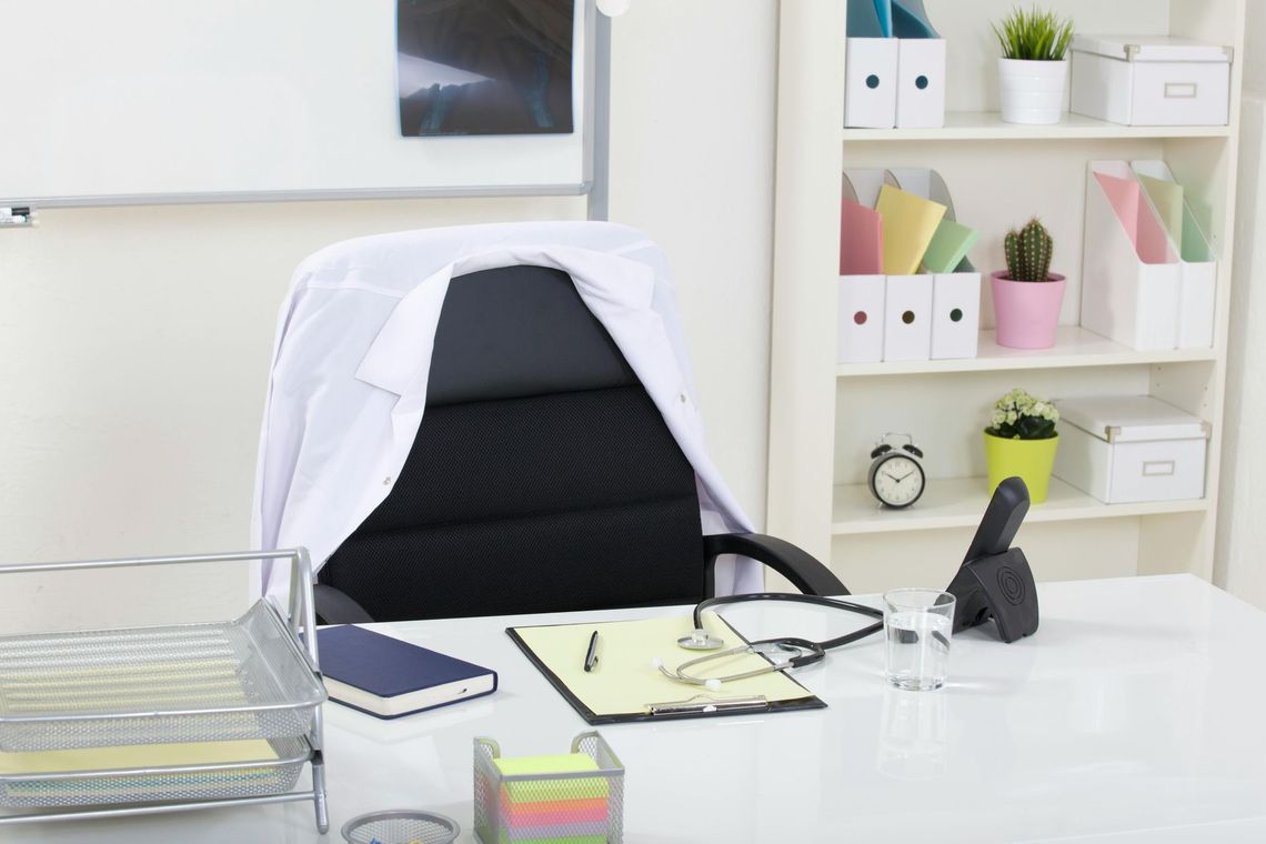 Bright office desk with black chair, white jacket, papers, laptop, and shelves with storage boxes.