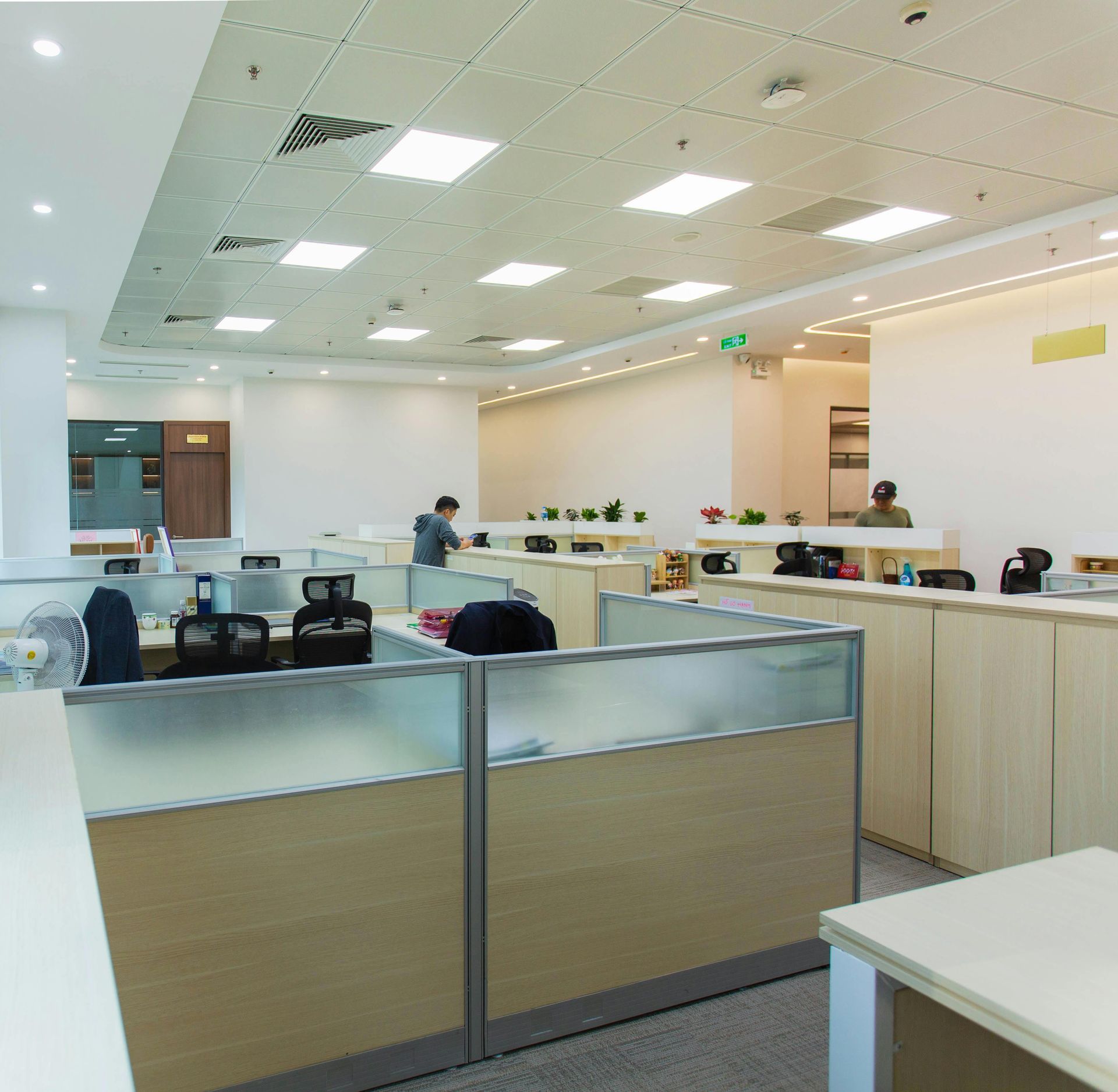 Modern open-plan office with beige cubicles, desks, and a few people working under bright ceiling lights