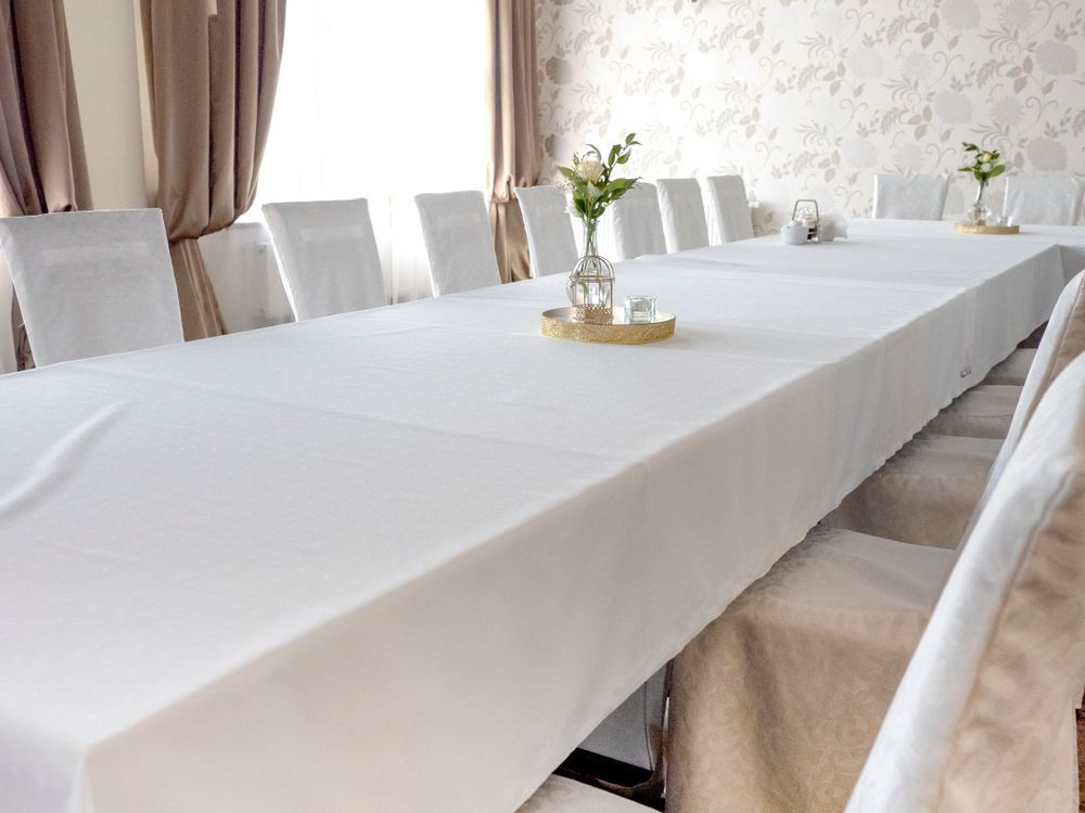 Long white banquet table with white chair covers and small floral centerpieces in a bright room