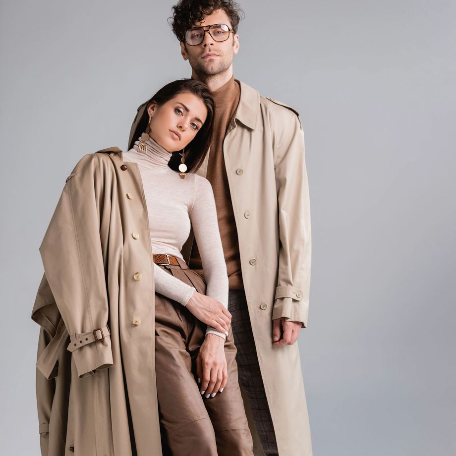 Due persone in posa insieme davanti a uno sfondo grigio uniforme, entrambe con indosso un trench color cachi e una maglia a collo alto.