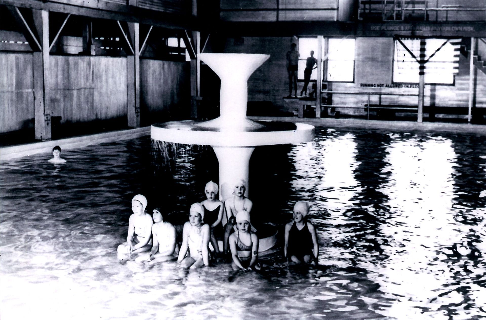 People standing in an indoor pool beneath a fountain in a dimly lit facility