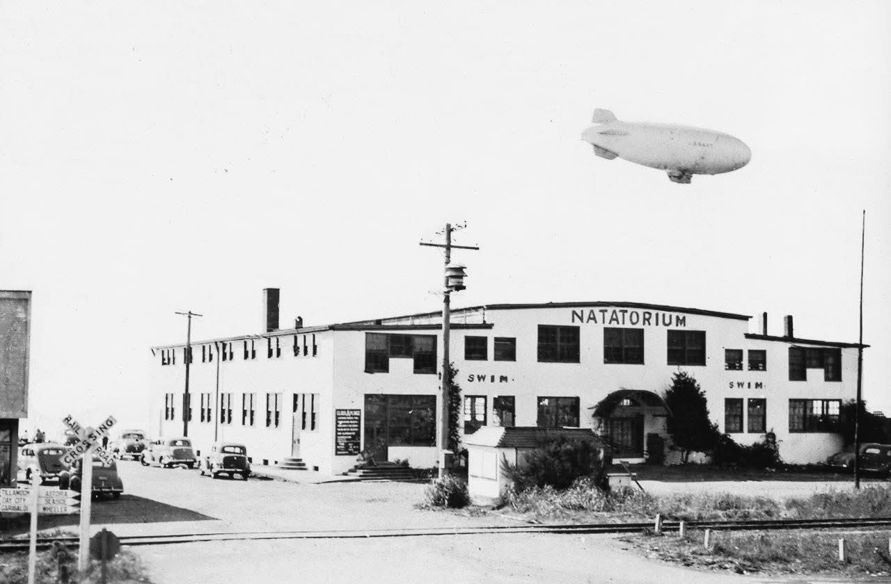 Black-and-white photo of a Natatorium building with a blimp flying overhead