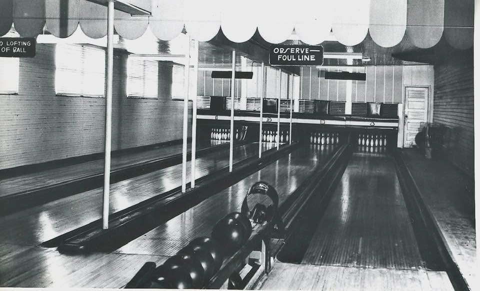 Black-and-white interior of a vintage bowling alley with lanes, pins, and overhead lights