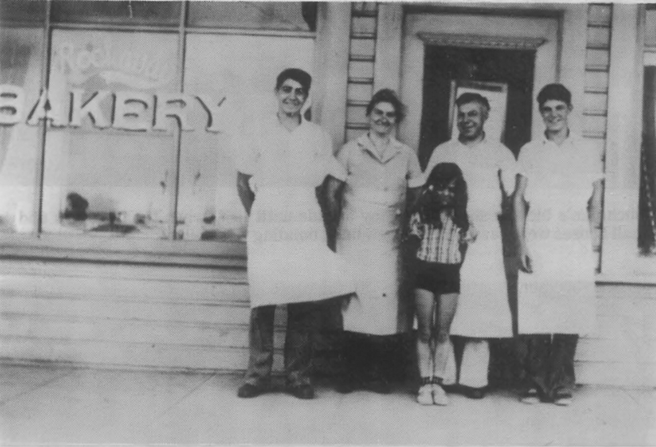 Four people standing outside a bakery storefront in aprons and uniforms, a young girl standing center in front