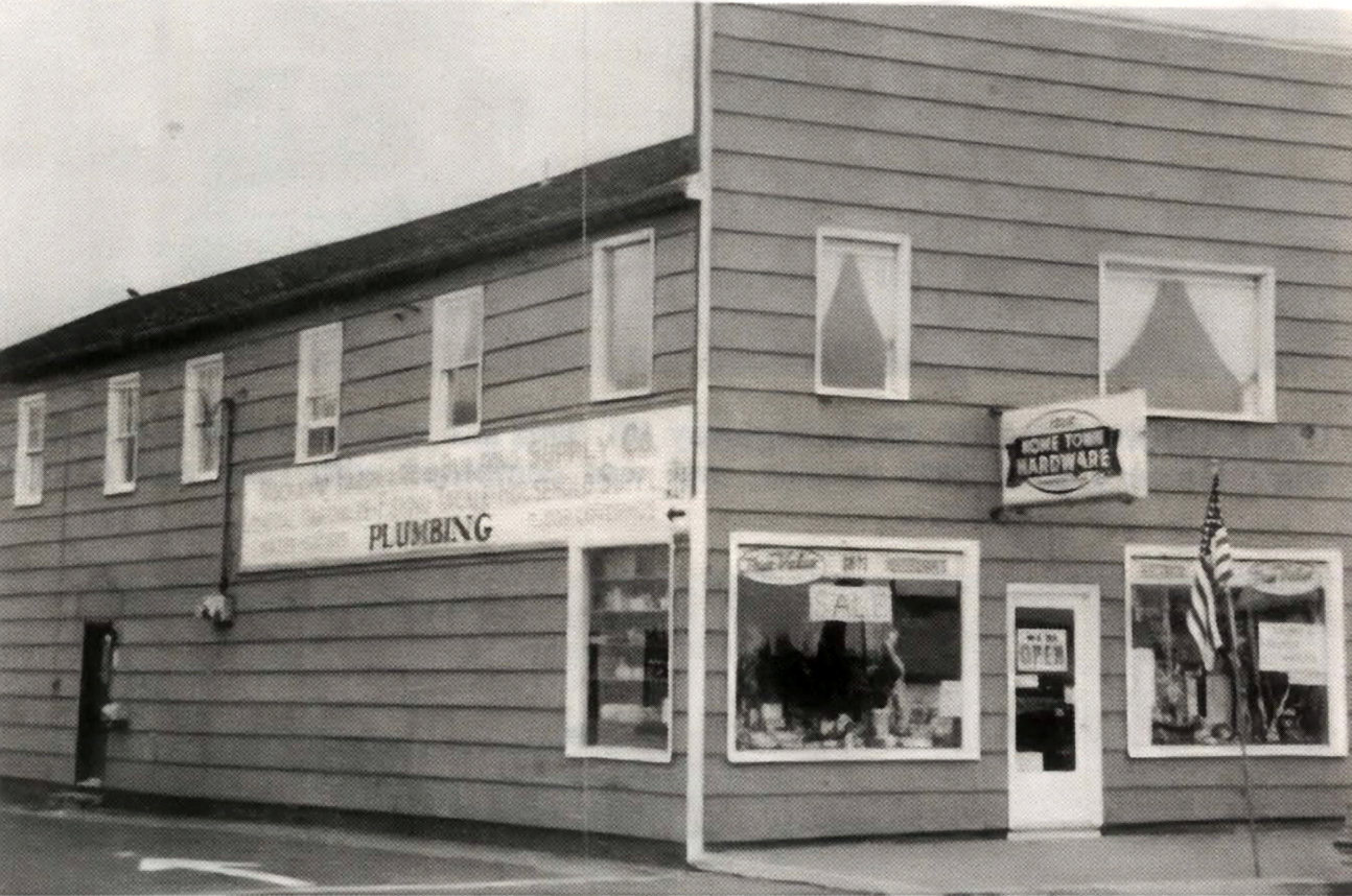 Black-and-white photo of a corner storefront with large display windows and a sign reading “PLUMBING”