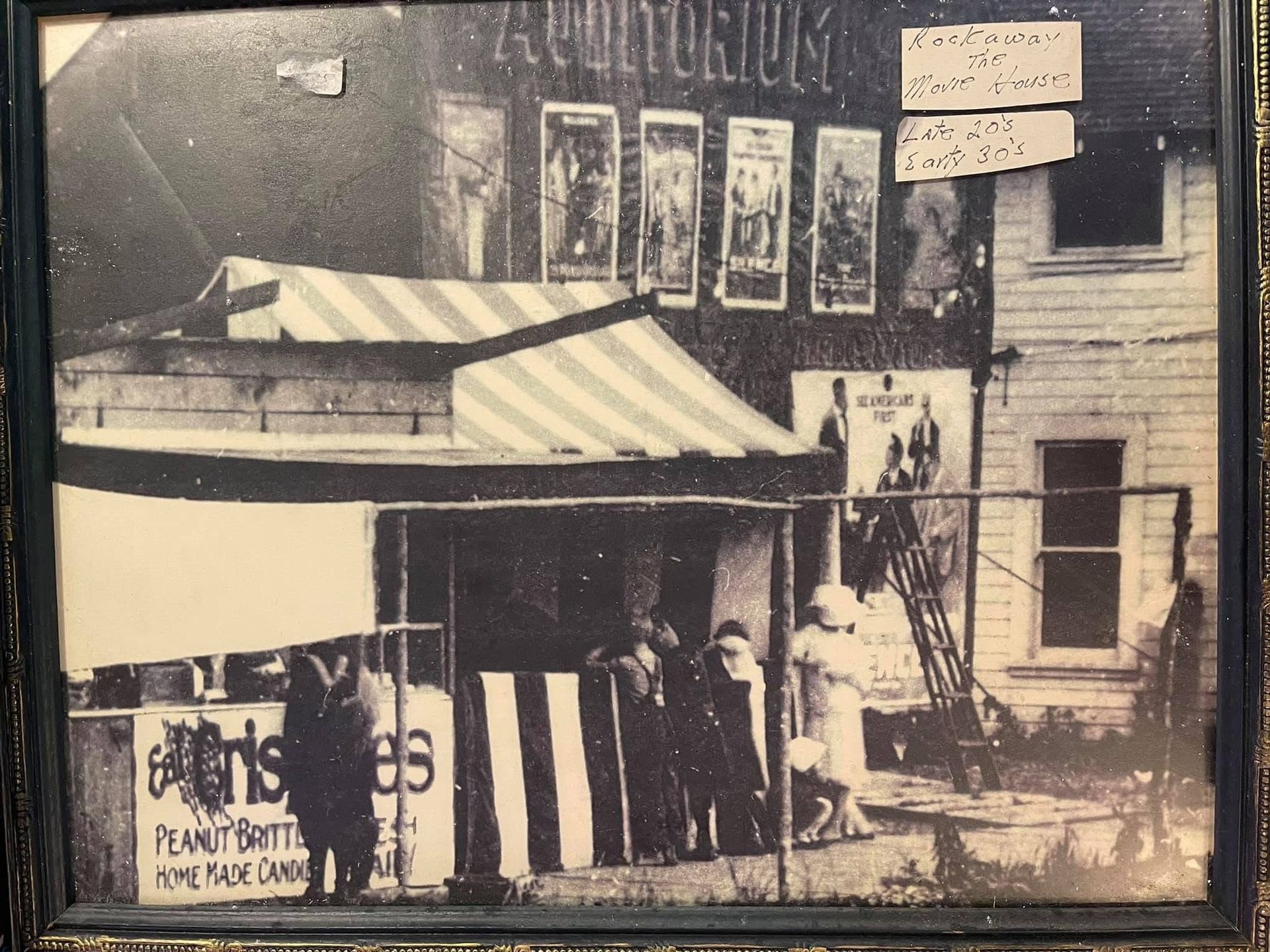 Old tent auditorium with a striped awning, posters on the wall, and people standing outside.