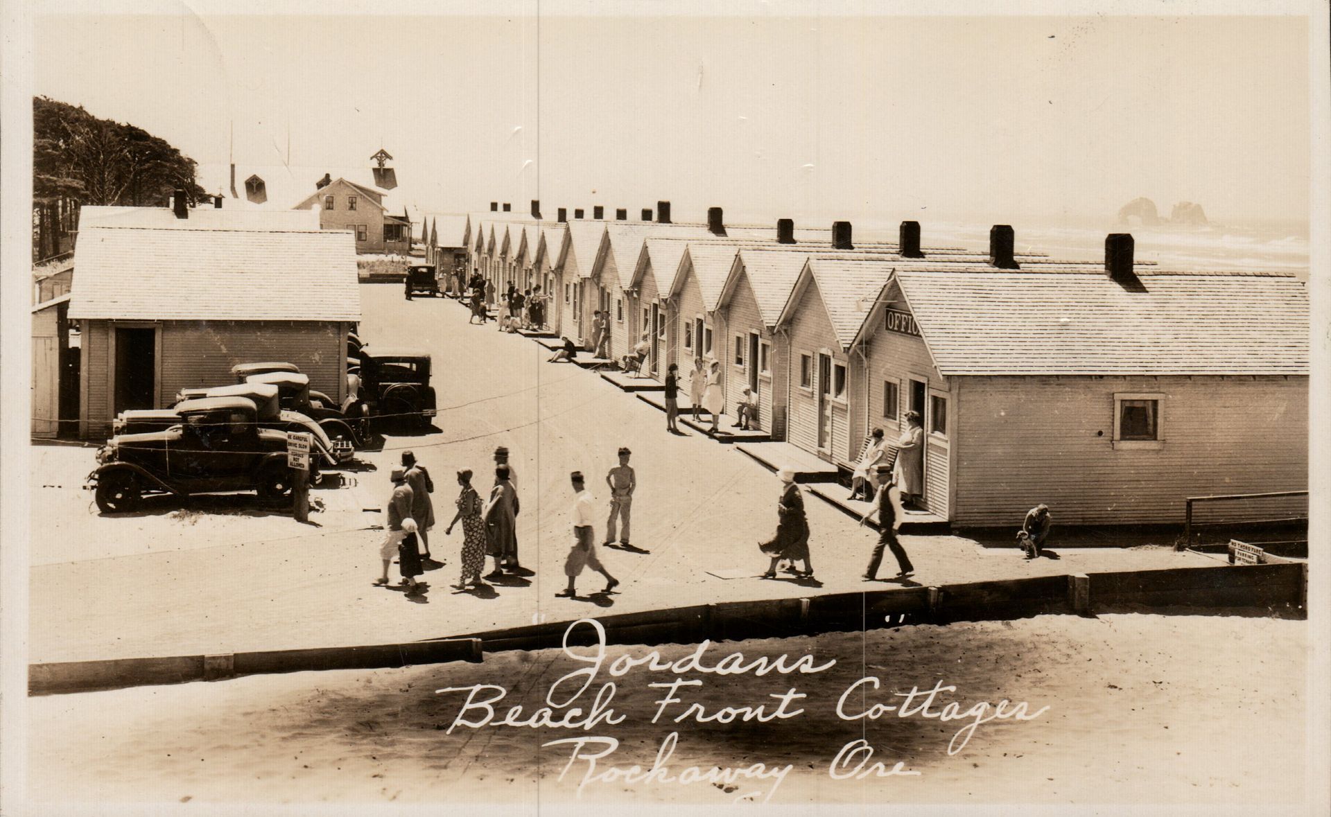 Sepia beach-front cottages in a row, with people walking along a path in front.