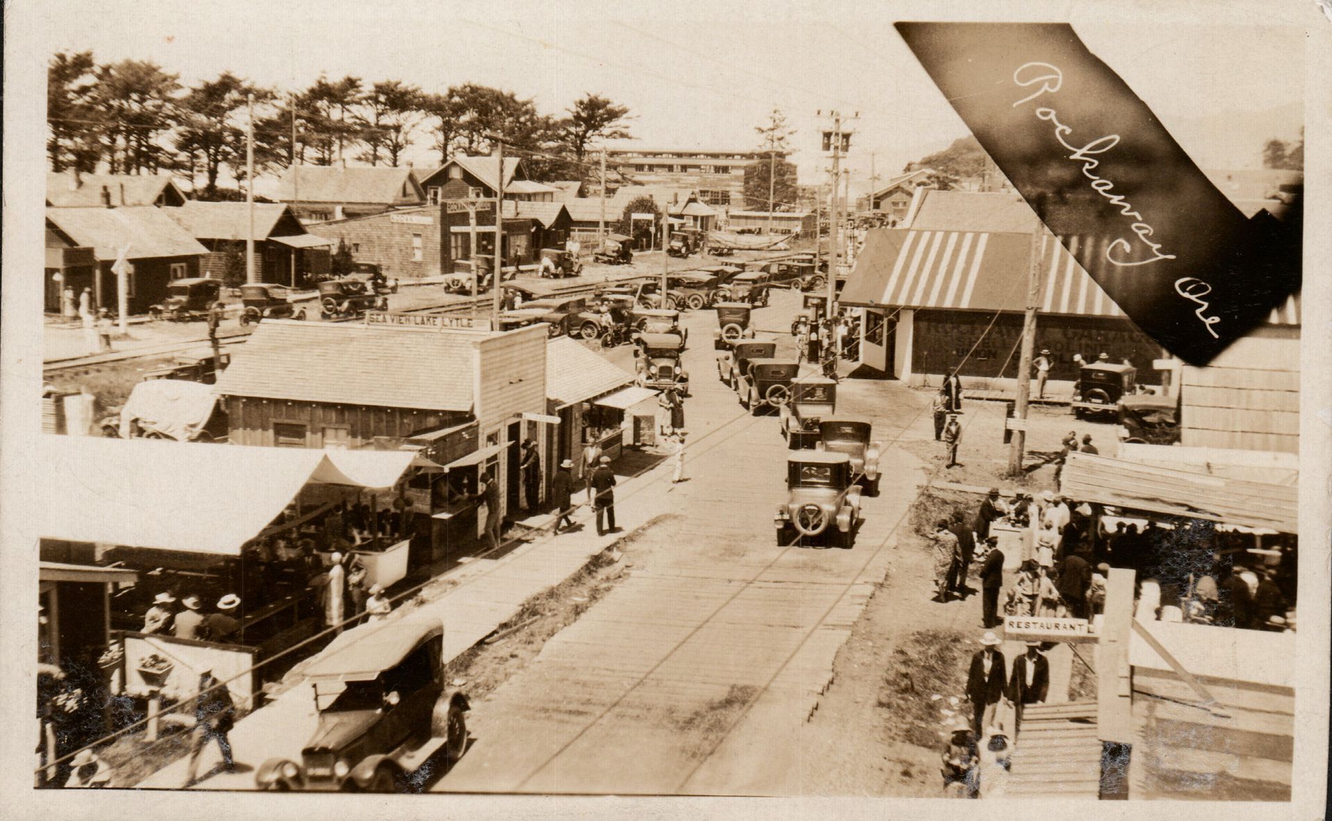 Vintage sepia street scene with early cars, storefronts, and pedestrians along a small-town main road