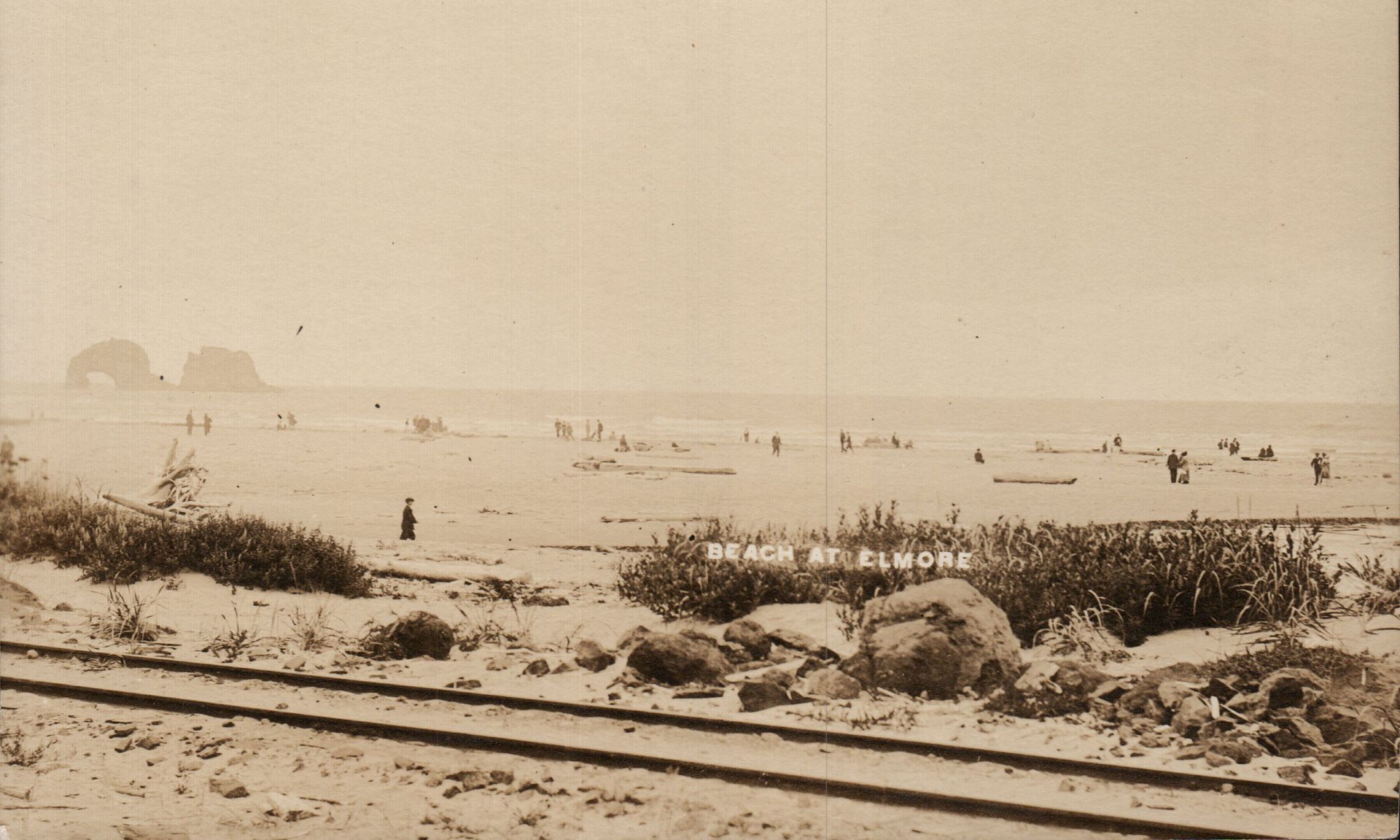 Sepia beach scene with people on the shore, distant sailboats, and railway tracks in the foreground