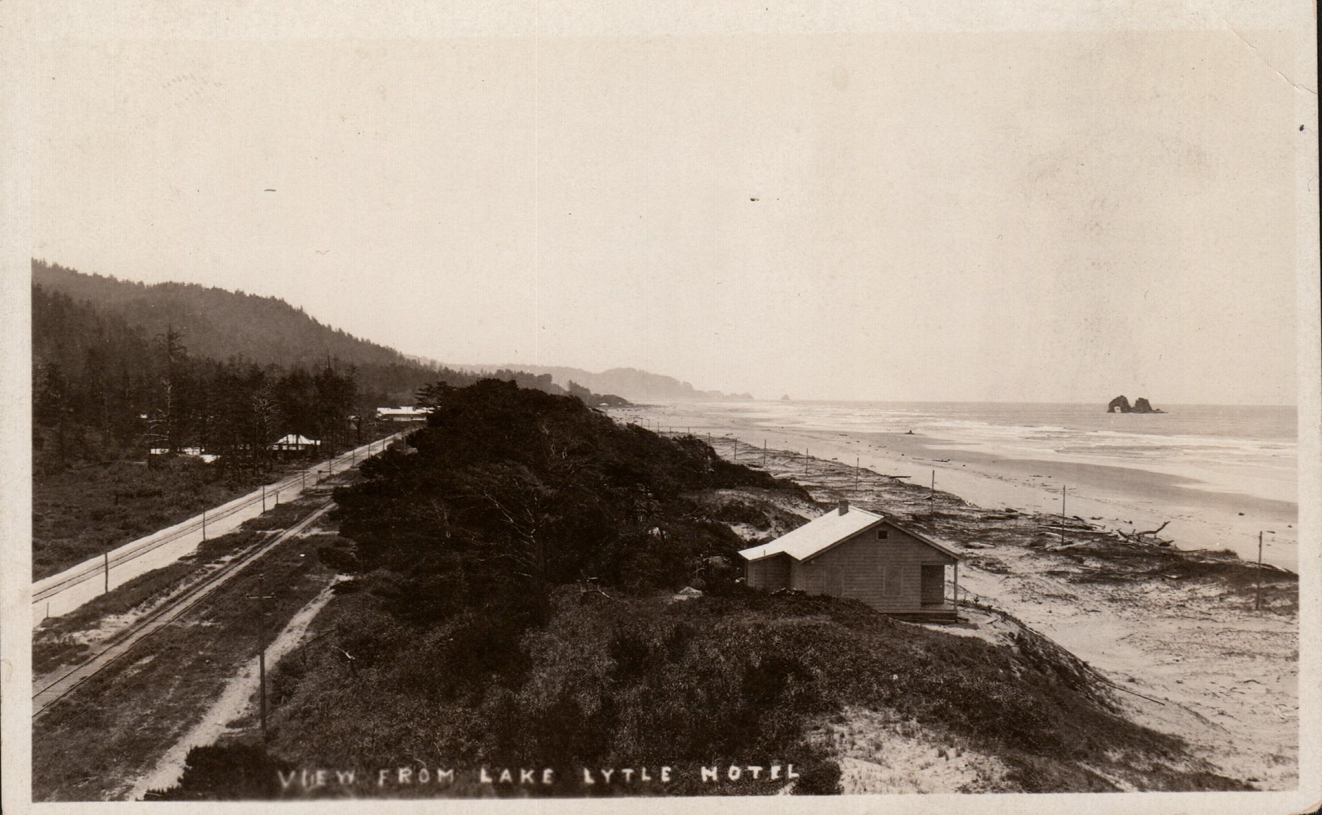 Black-and-white coastal view of a beach and road beside cliffs, with waves and distant rocks.