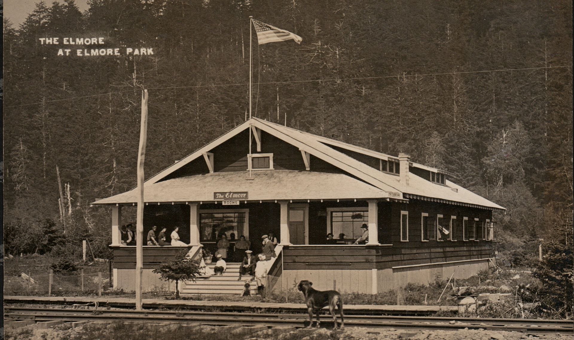 Sepia photo of a small train station with people on the platform and tracks in front.