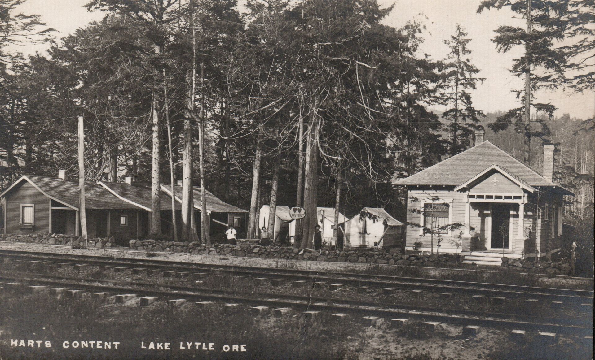 Historic lakeside resort buildings and cabins among tall trees, with railroad tracks in the foreground.